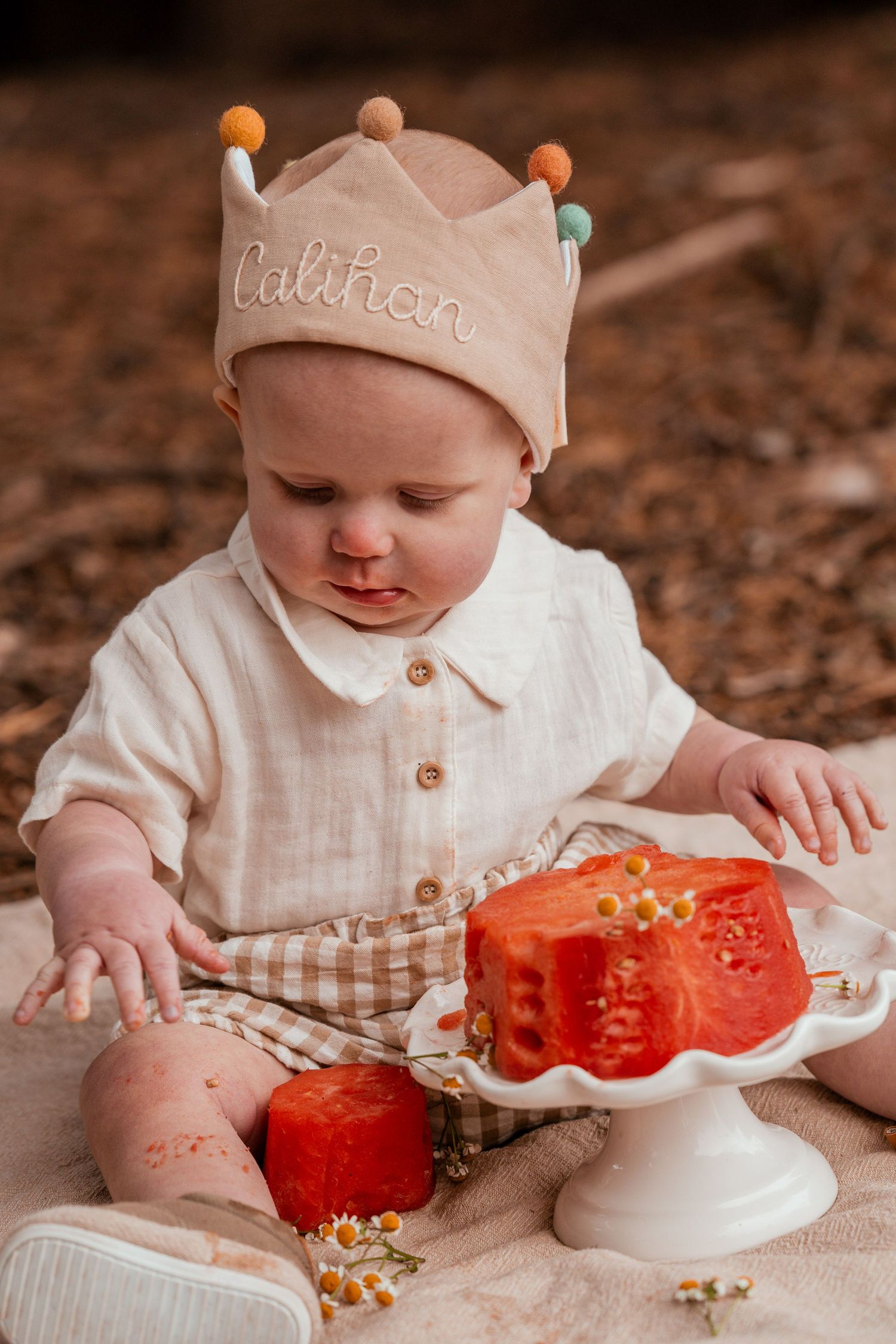 Baby wearing beige crown and white outfit explores red tomatoes on decorative white cake stand during outdoor photo session.
