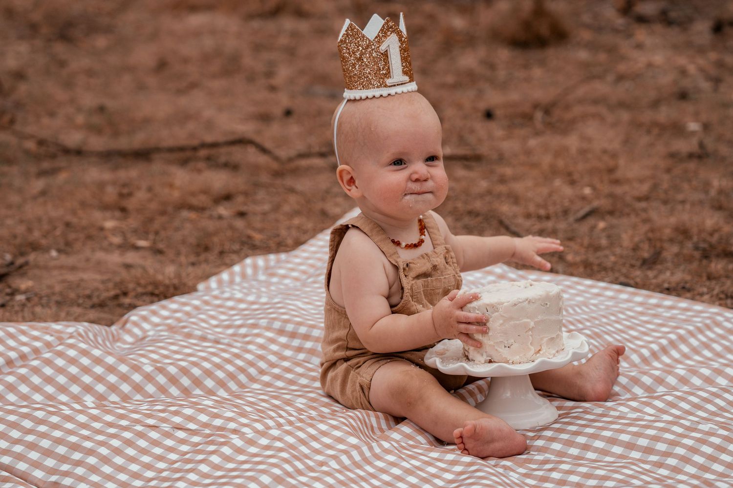 A baby in tan clothing sits on a checkered blanket outdoors while playing with a white plate.