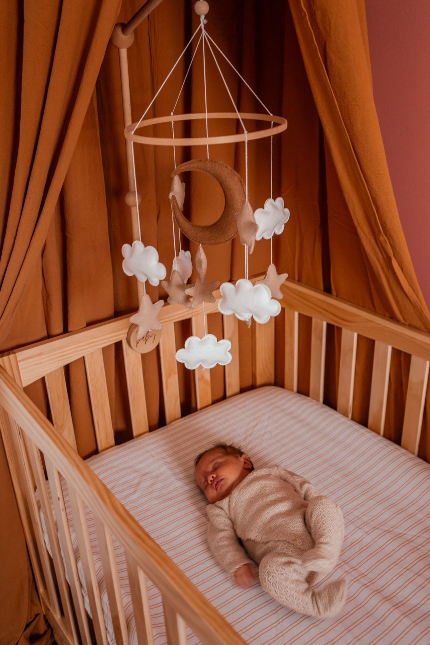 A wooden crib with a cloud and moon mobile hanging above while a baby sleeps on striped bedding.