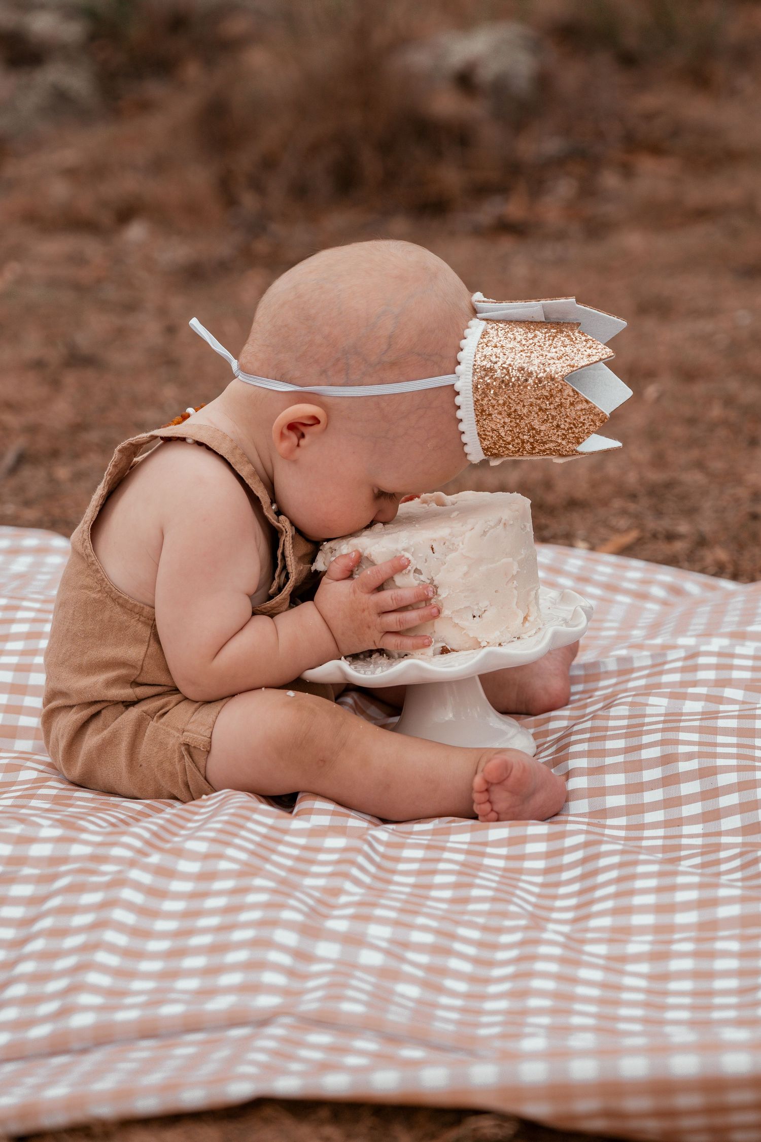 Baby wearing a glitter crown and brown romper sits on checkered blanket eating birthday cake.