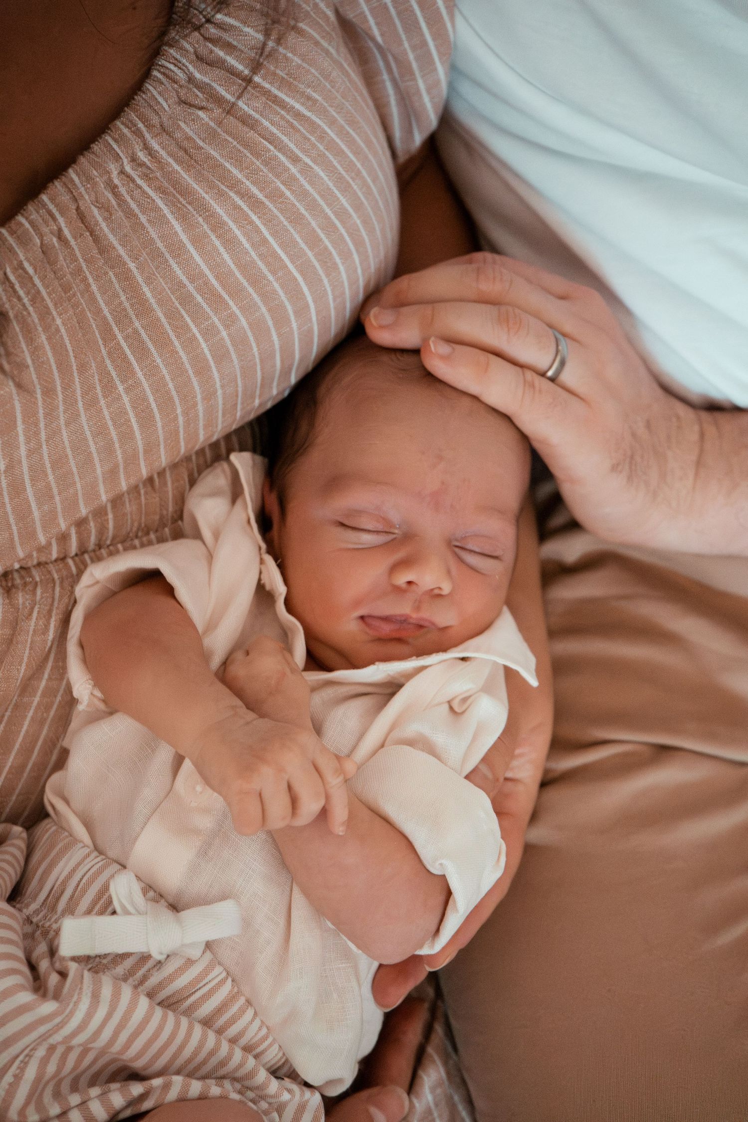 Sleeping newborn baby in cream outfit nestled between parents wearing light colored clothing.