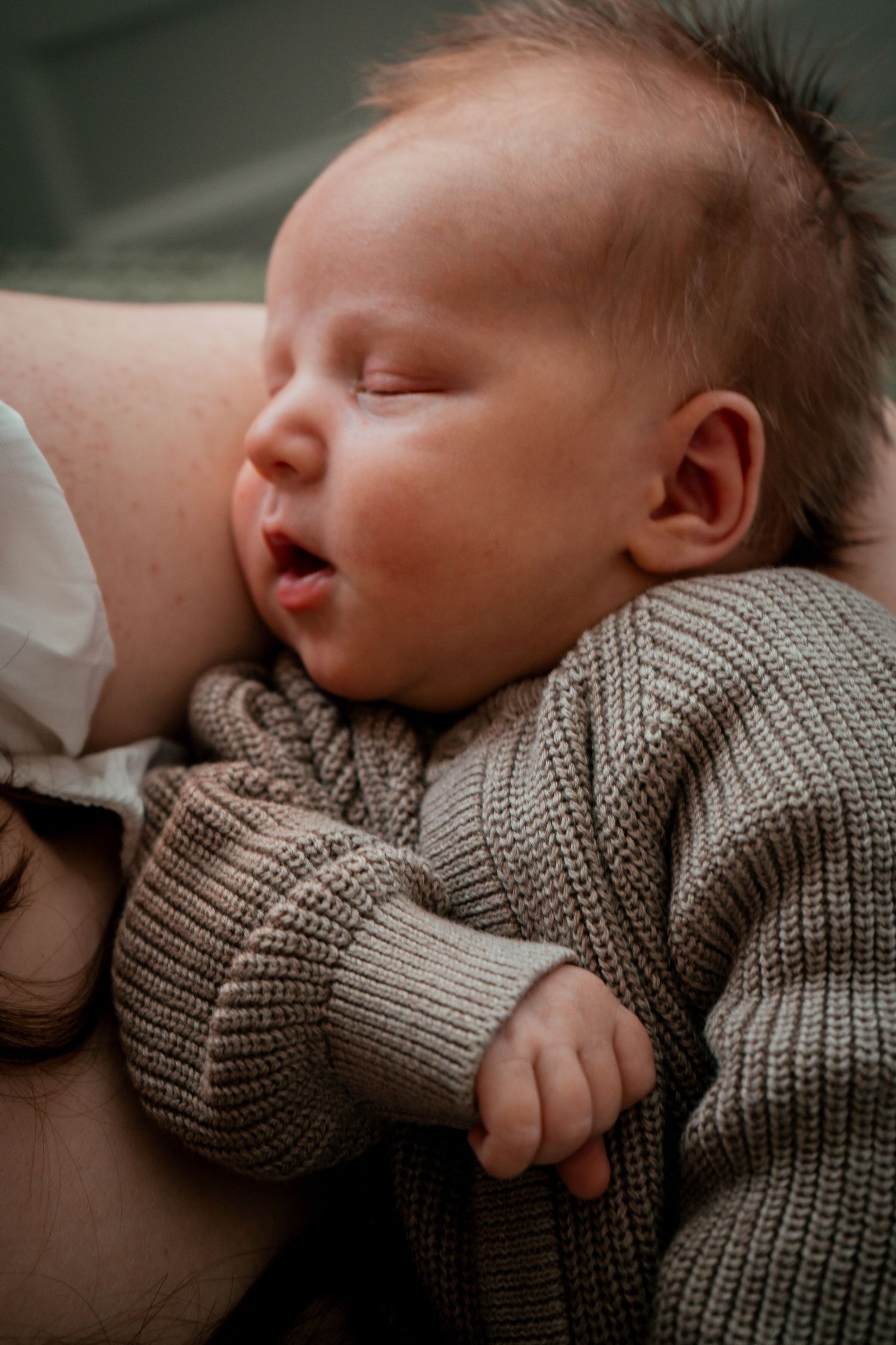 A sleeping newborn baby wearing a cozy gray knit sweater rests peacefully on a soft surface.