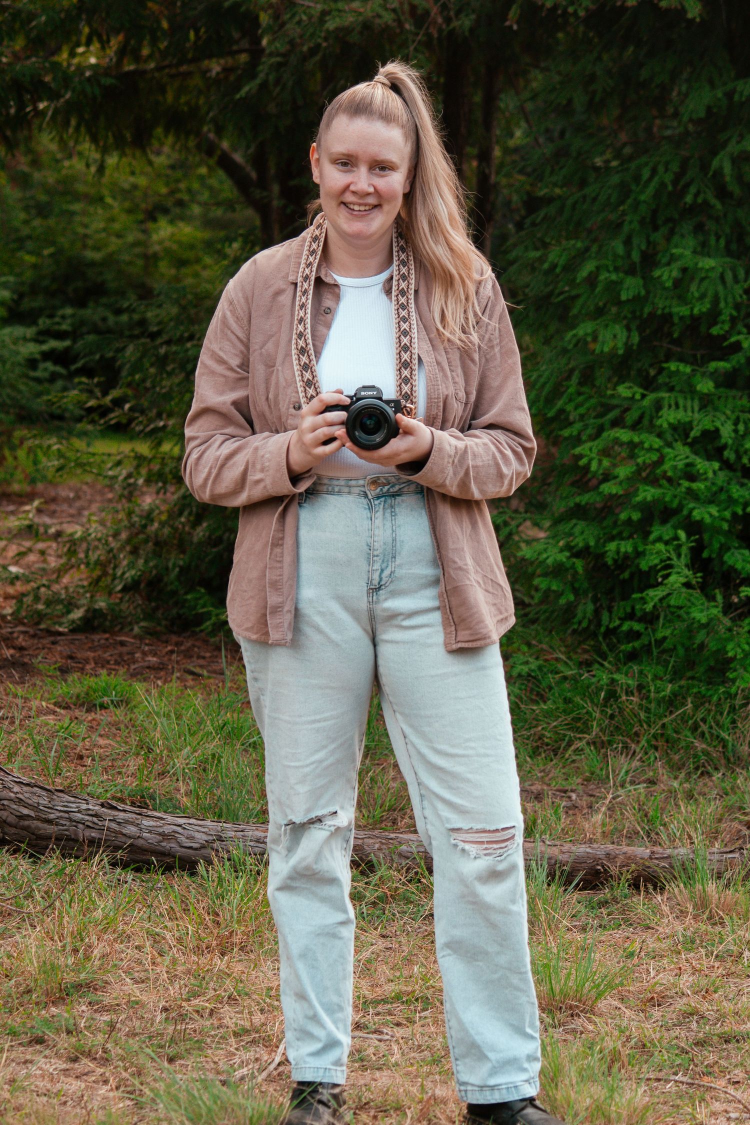 Someone poses with a camera while wearing a brown jacket and distressed jeans in a forest setting.