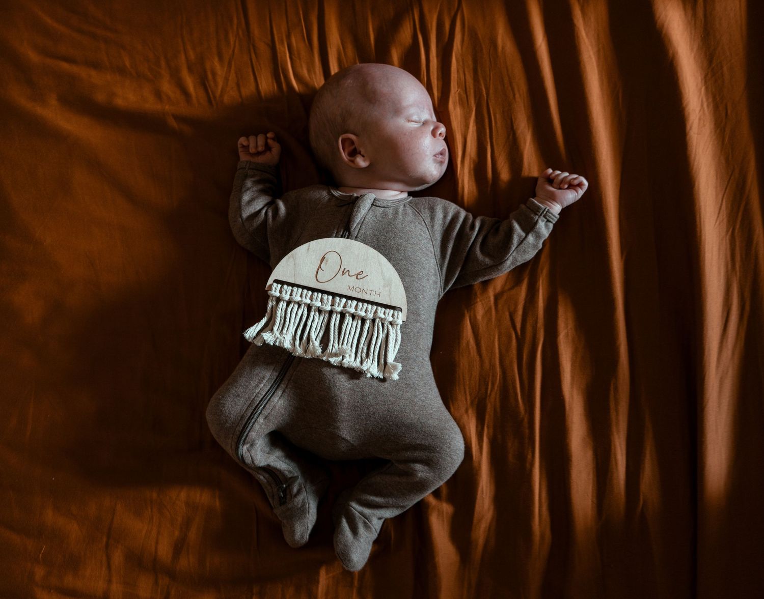 Sleeping in grey romper with fringe detail on brown fabric background.