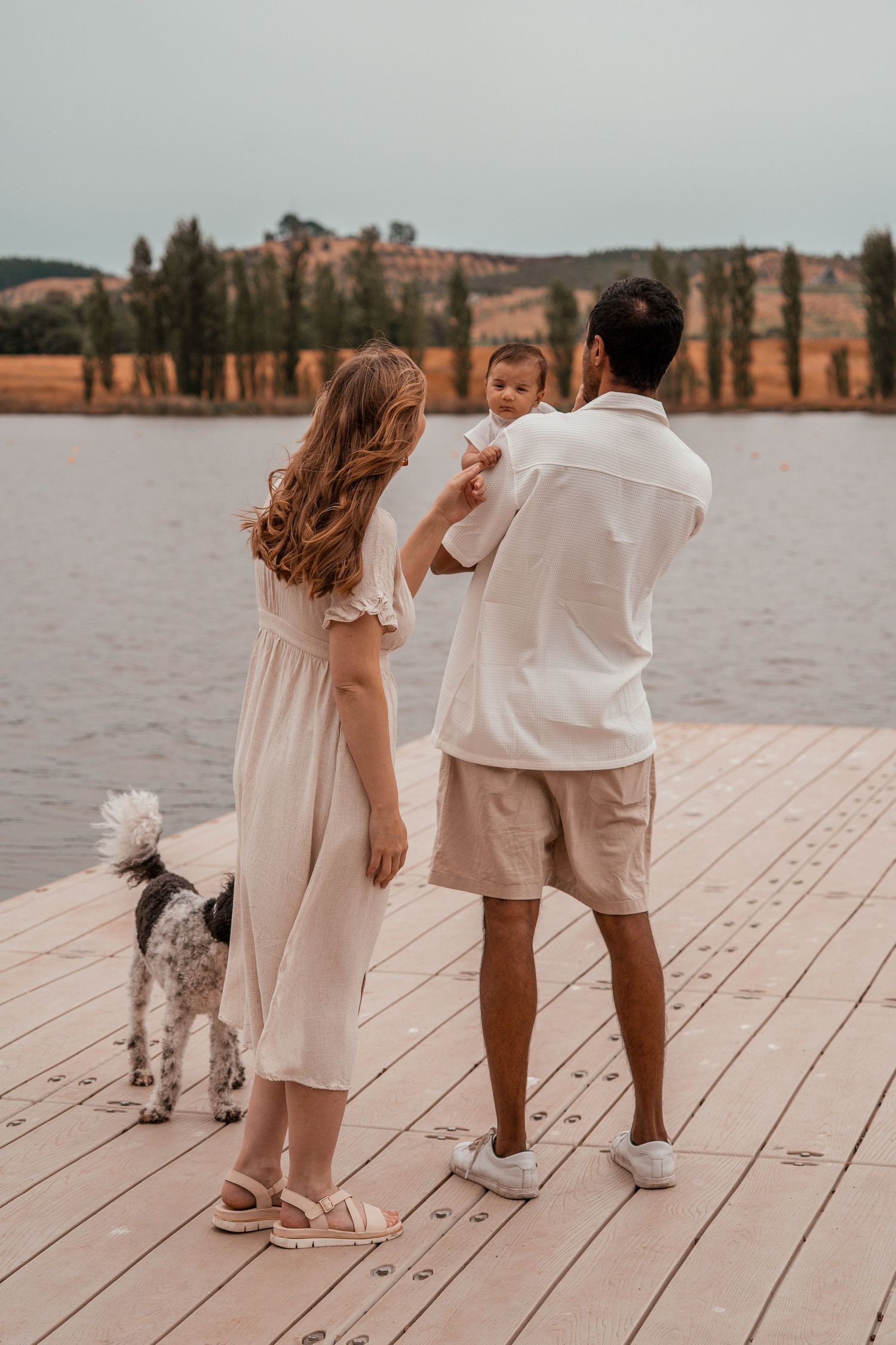 A family and their small dog walk together on a wooden dock by a serene lake during sunset.