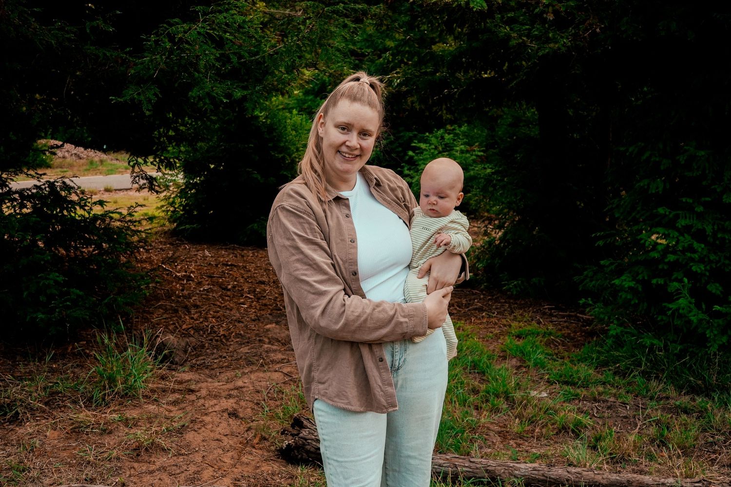 Casual outdoor portrait of parent and baby against green forest backdrop.