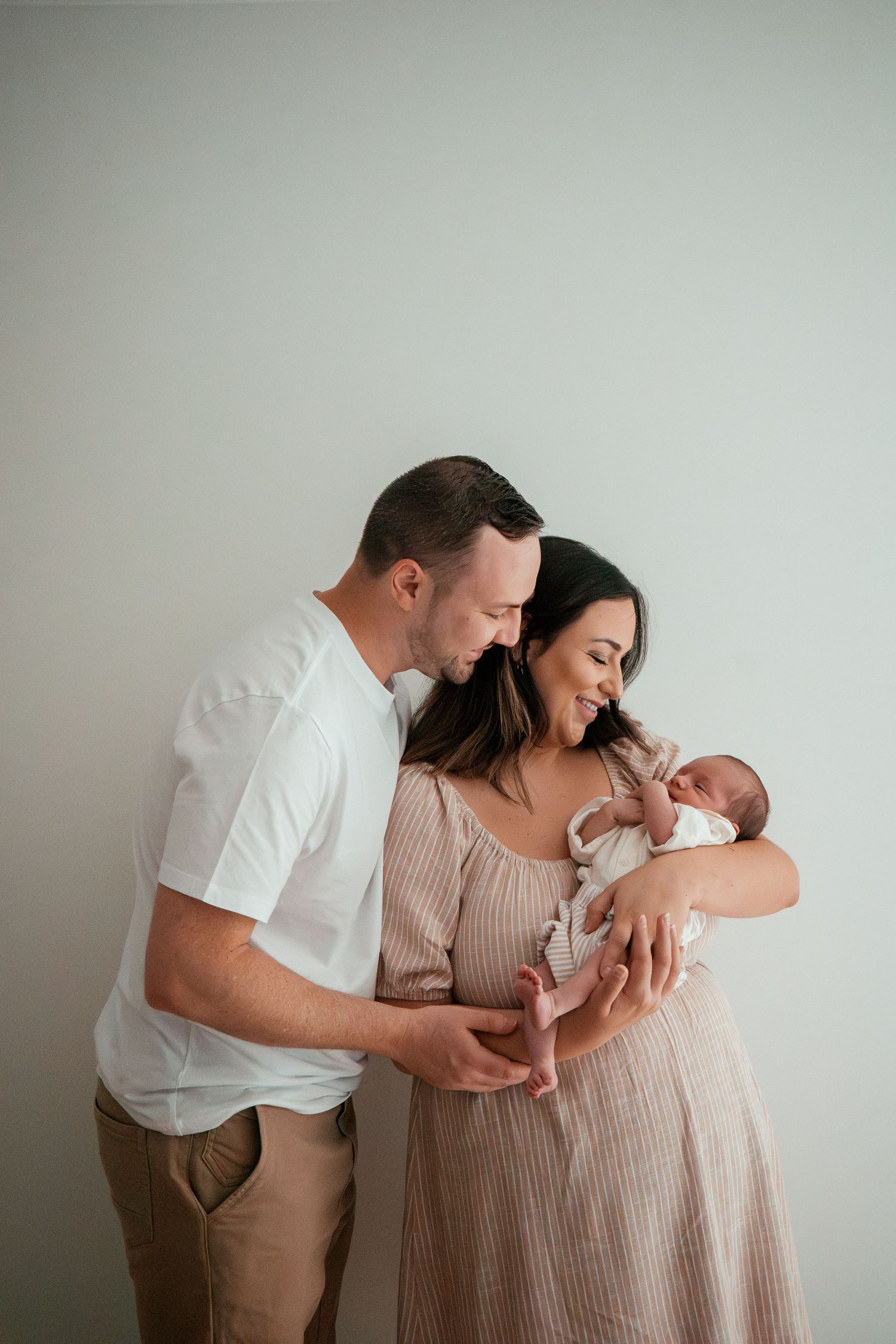 A couple in neutral tones sharing tender moments with their newborn baby in a minimalist studio setting.