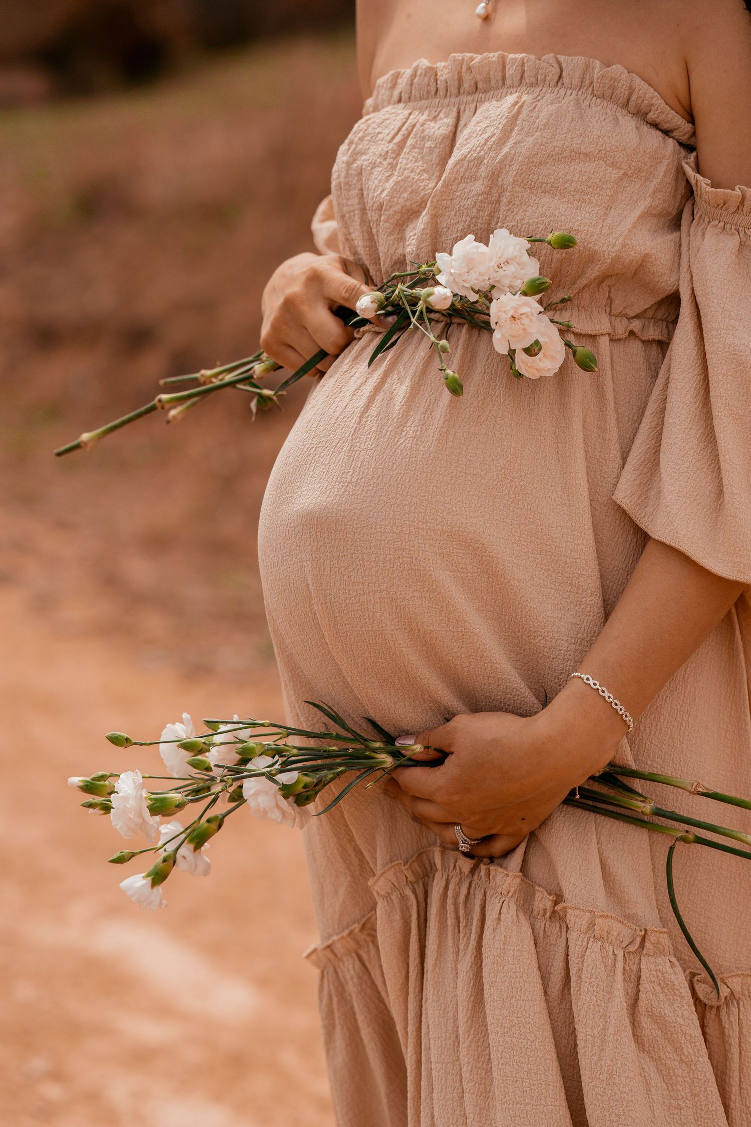 Pregnant woman in beige off-shoulder dress holding delicate white flowers against an earthy background.