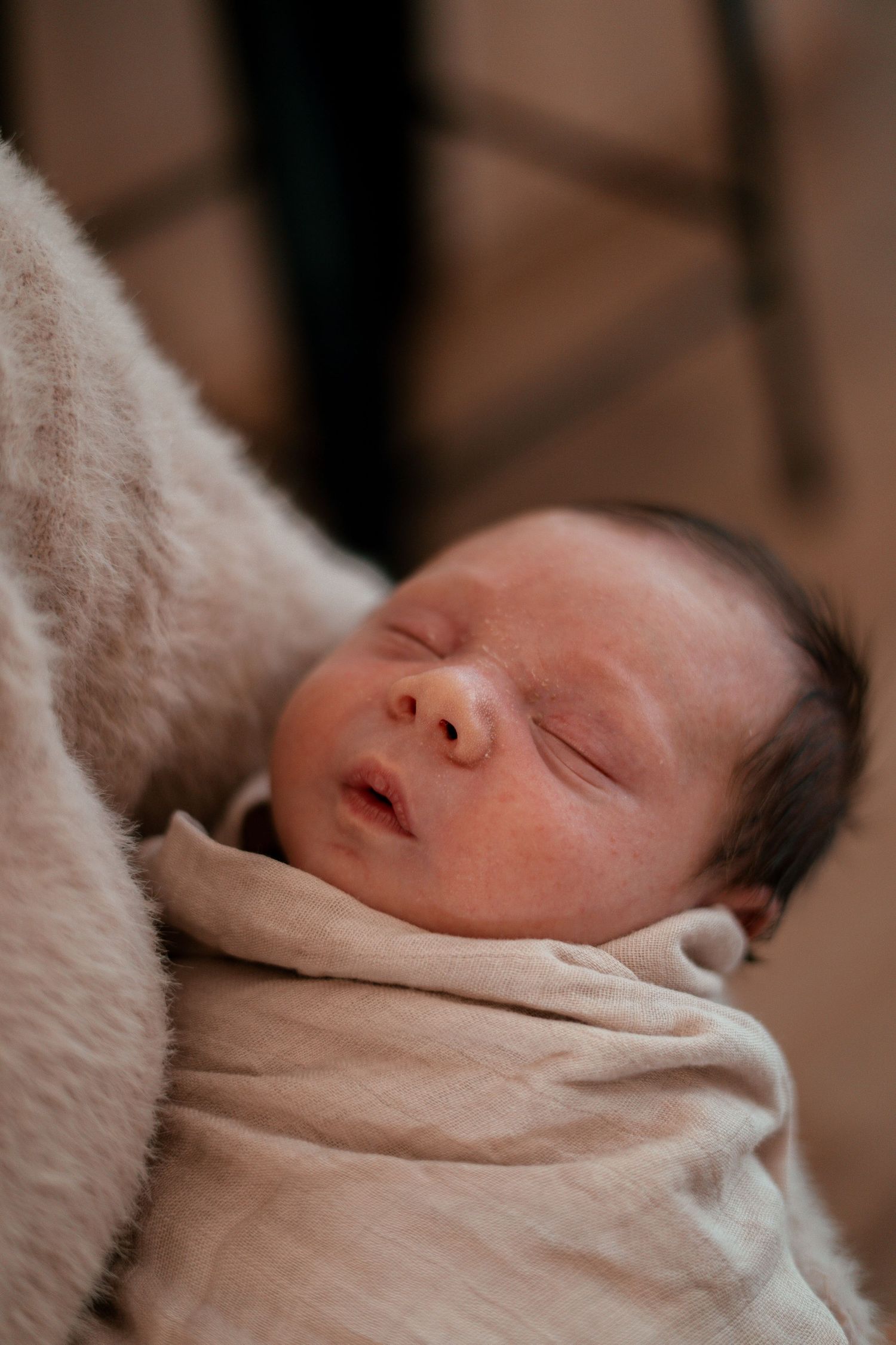 A sleeping newborn baby swaddled in soft cream-colored blankets against a warm brown background.