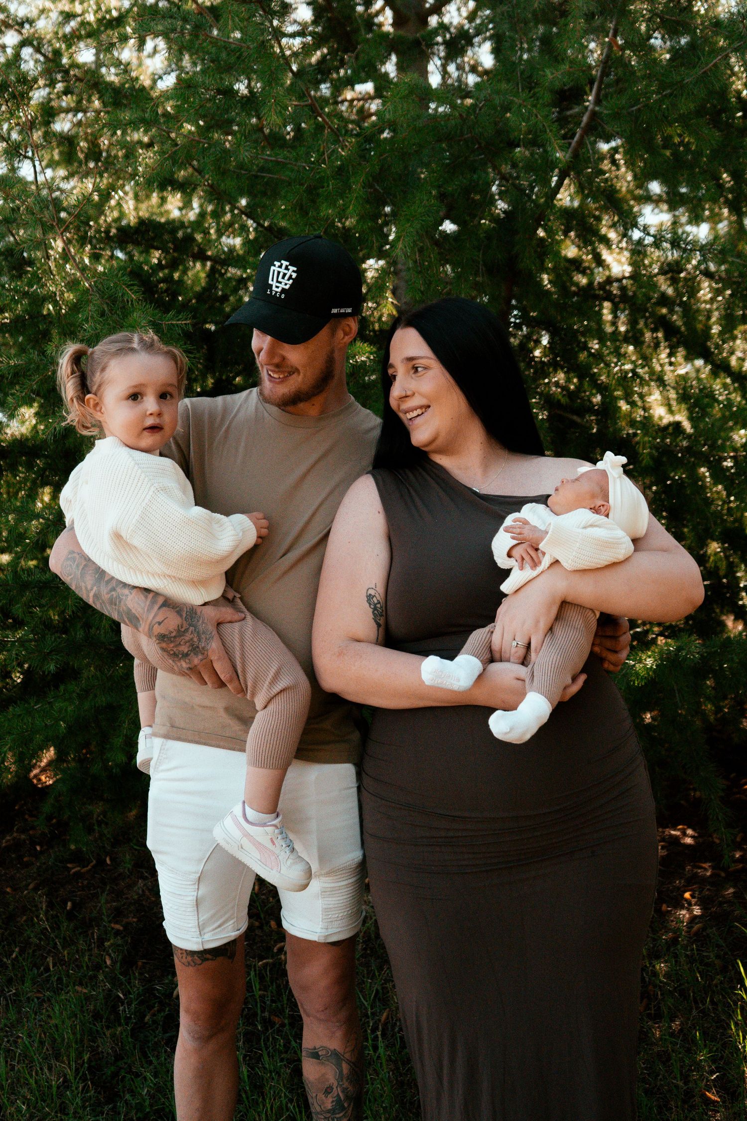 Family portrait outdoors with toddler and newborn against green foliage background.