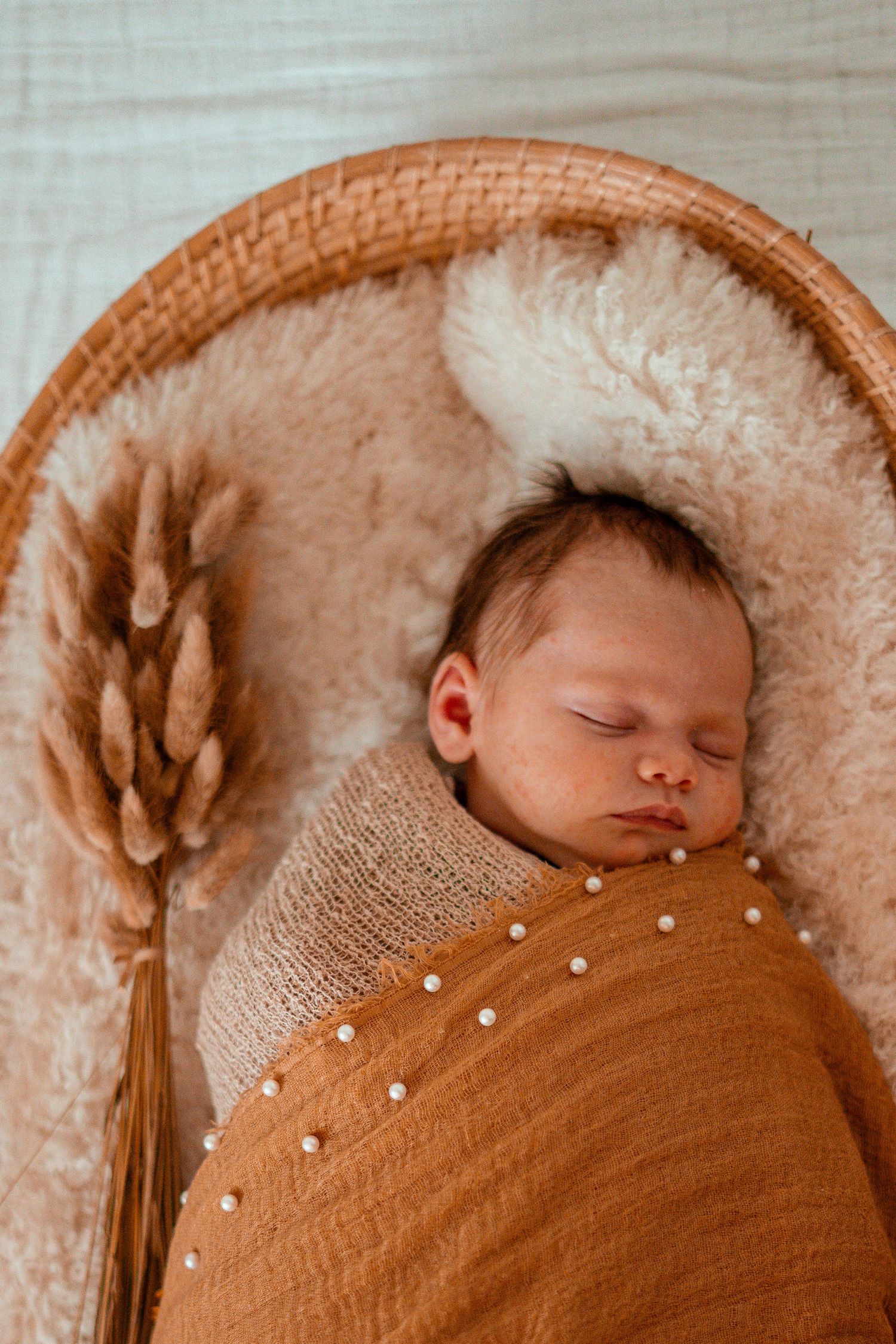 A sleeping baby wrapped in a brown knit blanket with pearl details lies on a cream-colored cushion next to dried wheat.