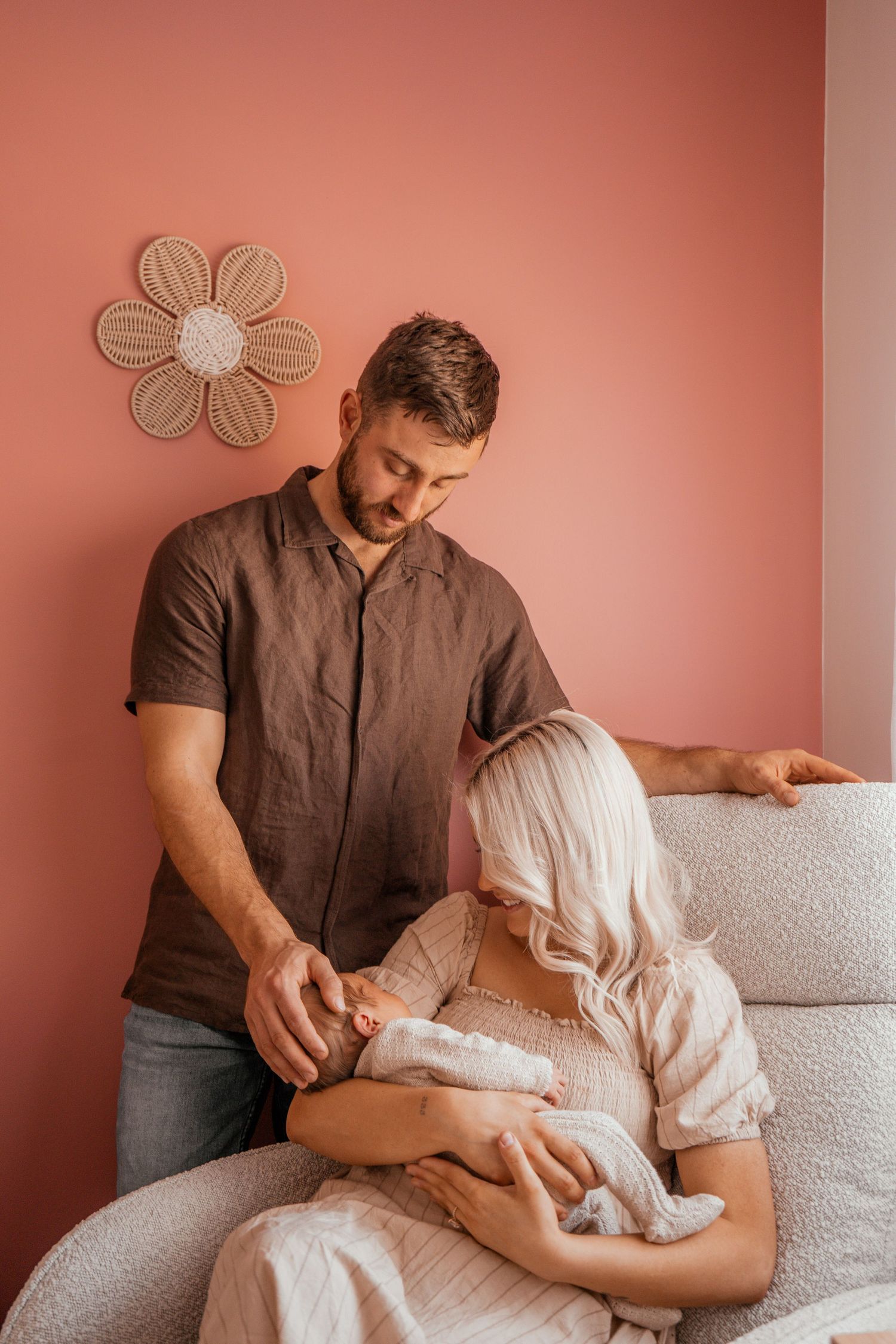 A couple shares an intimate moment with their newborn baby on a cozy couch against a pink wall with floral decor.