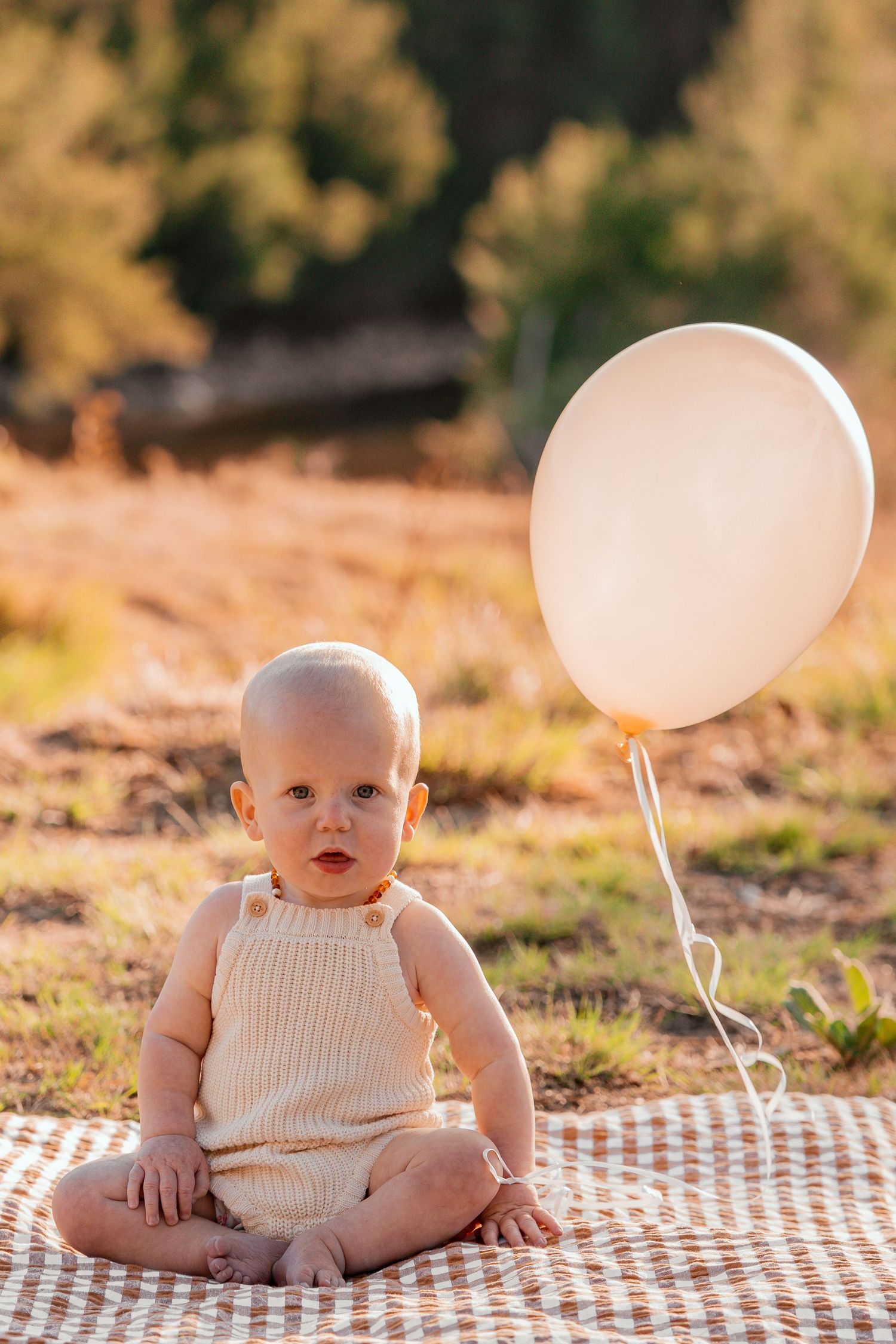 A baby in a white romper sits on a checkered blanket outdoors with pink balloons in golden sunlight.