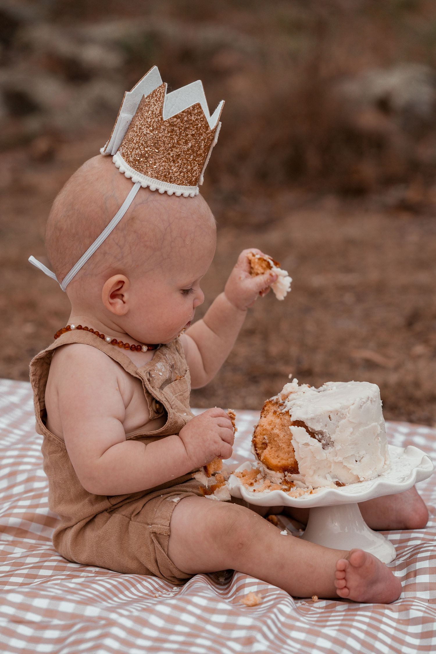 Baby in a gold crown sits on checkered blanket enjoying cake during first birthday cake smash photo session.