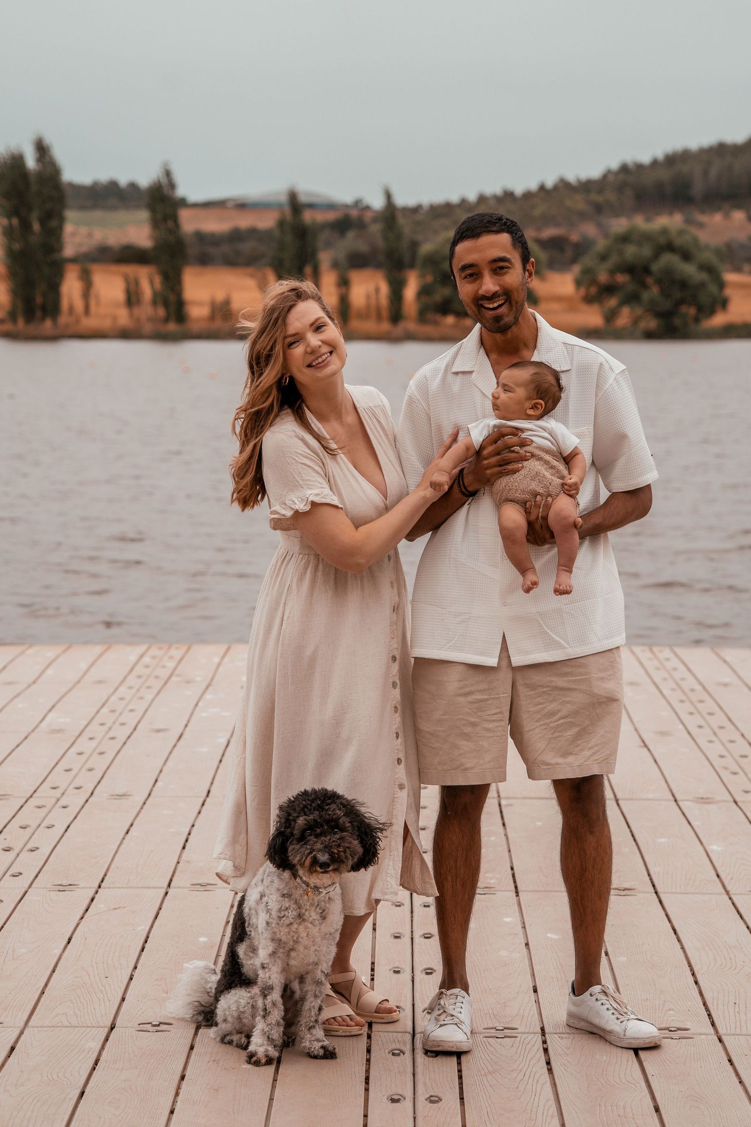 A happy family poses with their baby and dog on a wooden deck overlooking a scenic lake and mountain landscape.