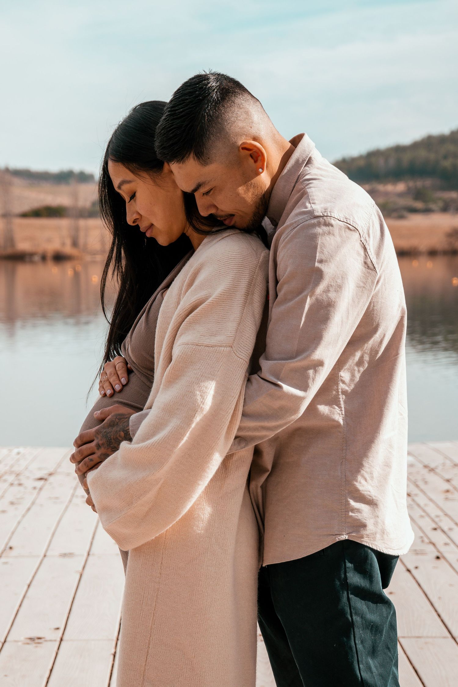 Tender romantic moment between couple embracing on wooden dock by lake during sunset in neutral colored clothing.
