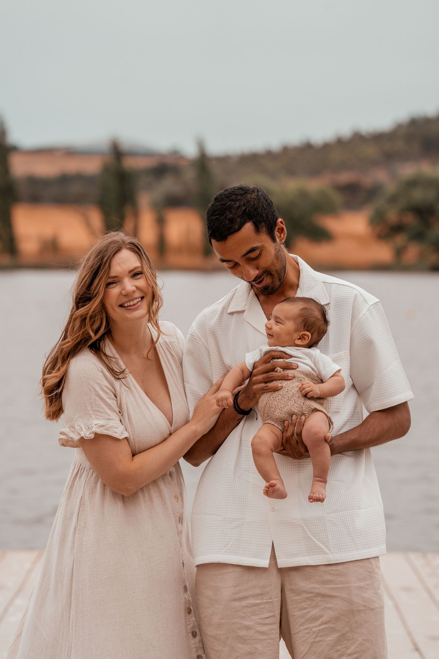 A couple in light-colored clothing shares joyful moments with their baby by a lake during a family photoshoot.