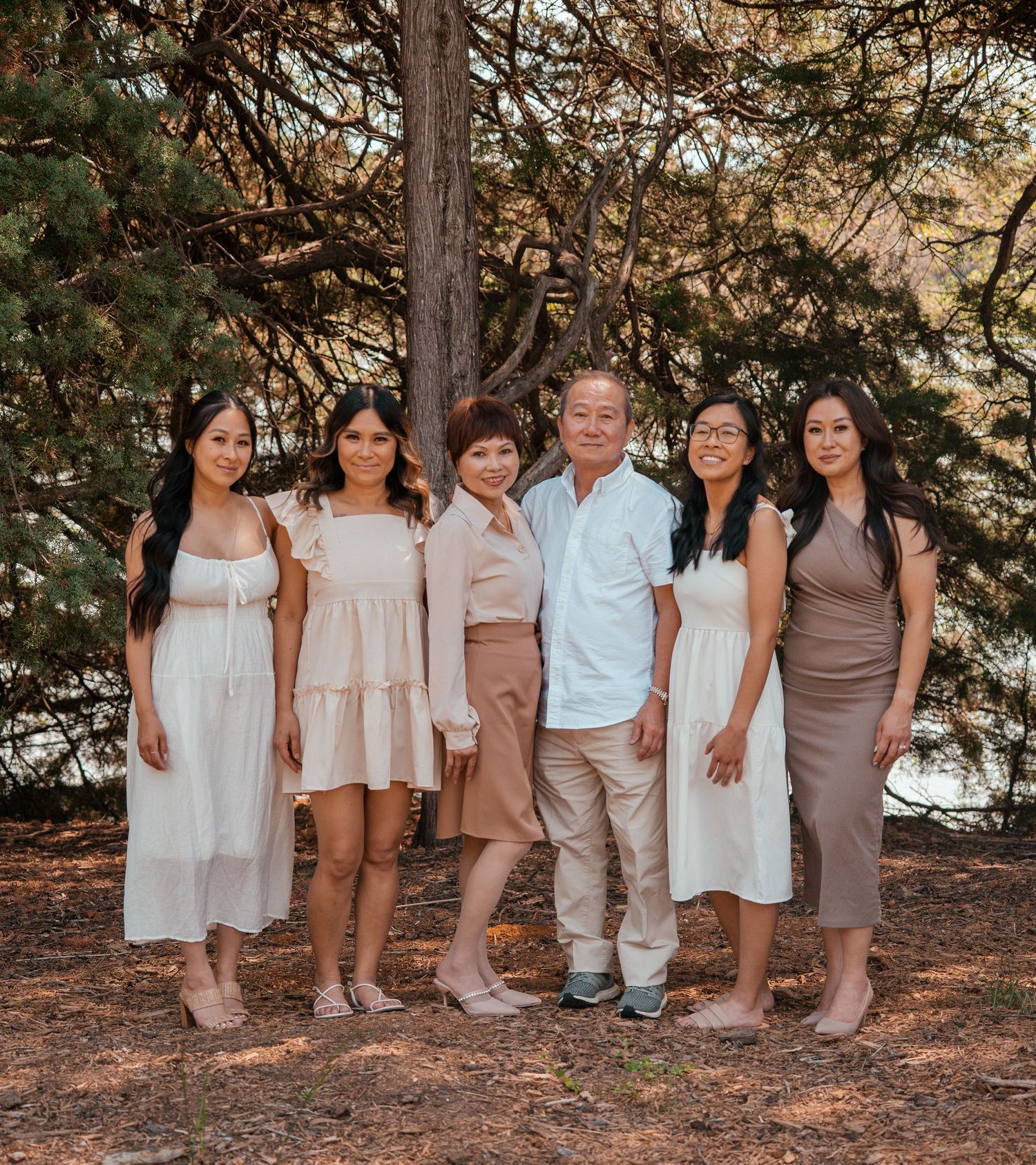A family group poses together under tall pine trees wearing coordinated white, brown and neutral toned outfits.