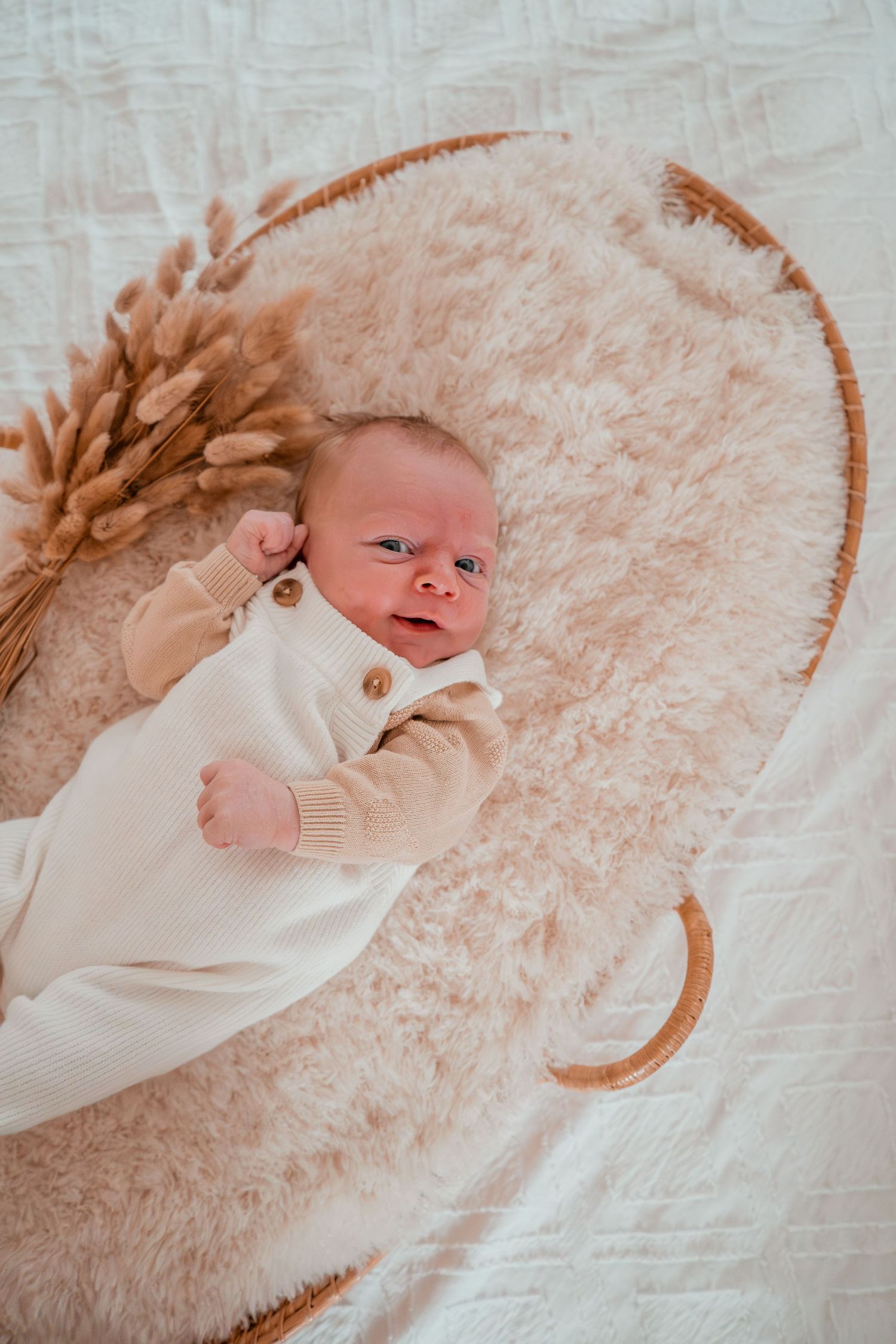 A newborn baby lies on soft white bedding with a curved wicker basket rim visible in the background.