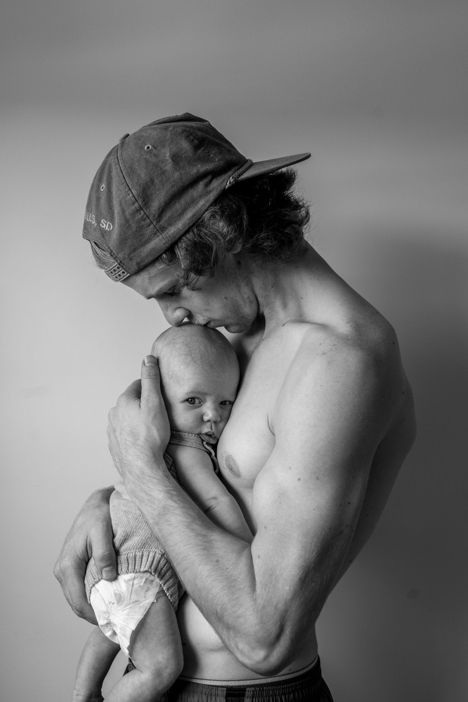 Intimate black and white portrait of shirtless parent holding infant against gray background.