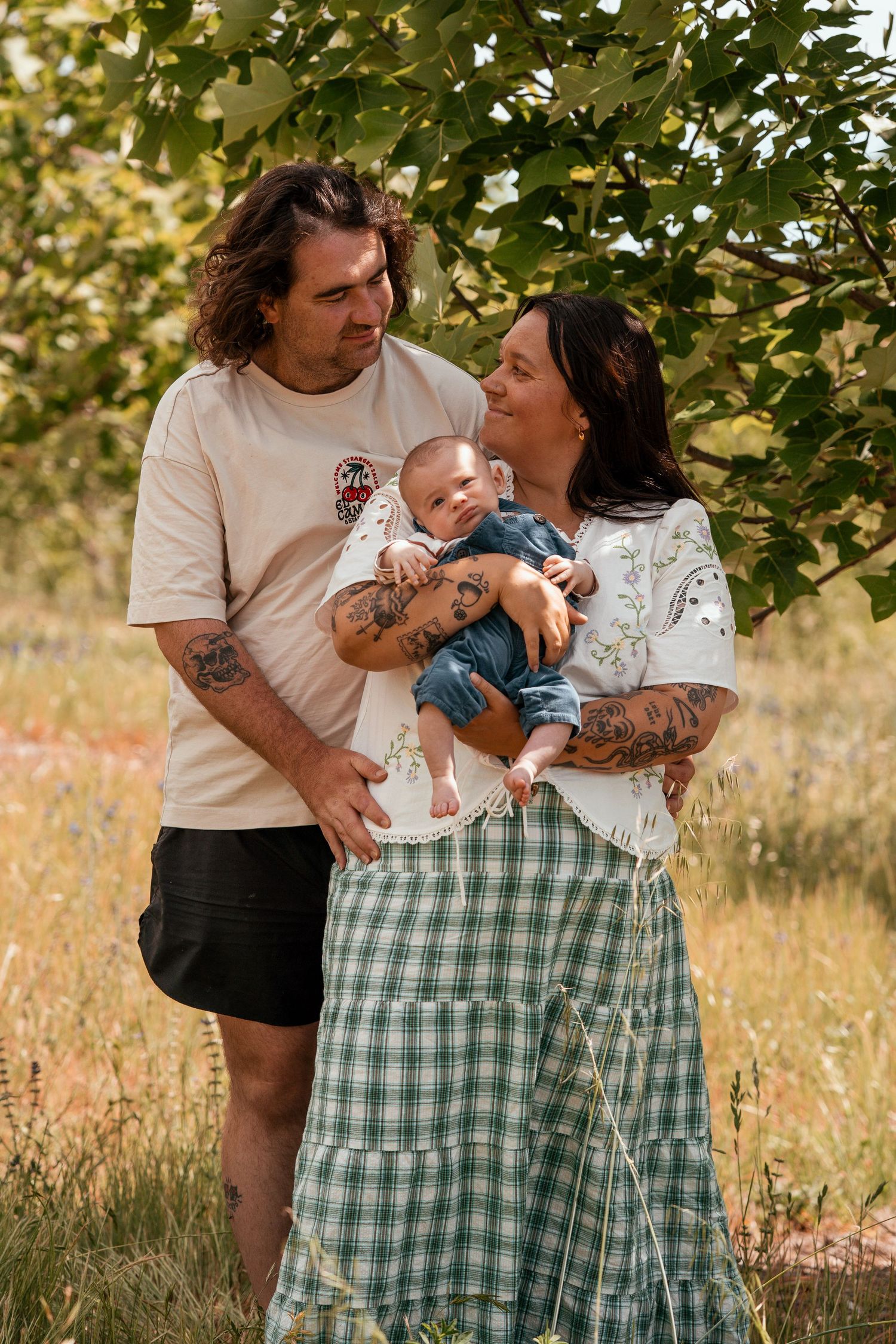 Family shares tender moments together in a sunny outdoor setting with green trees and wildflowers in the background.