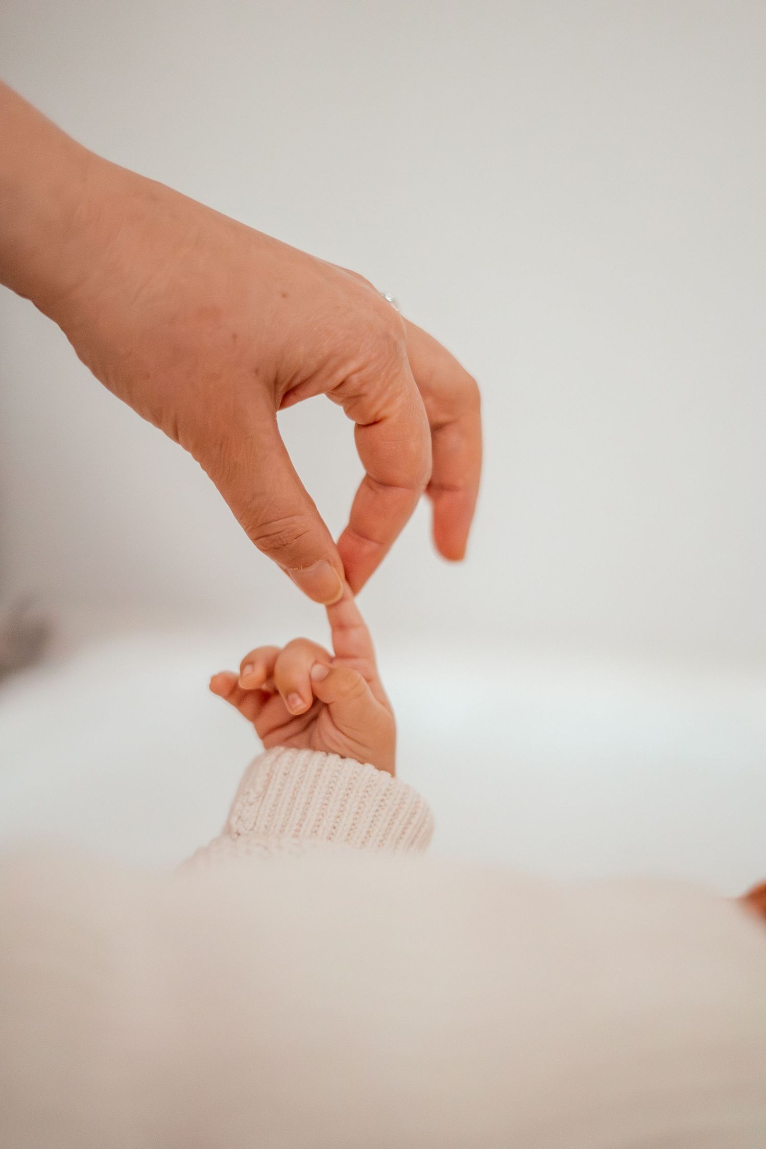 Adult hand gently touches tiny baby's hand wearing white knit sleeve in tender moment against light background.