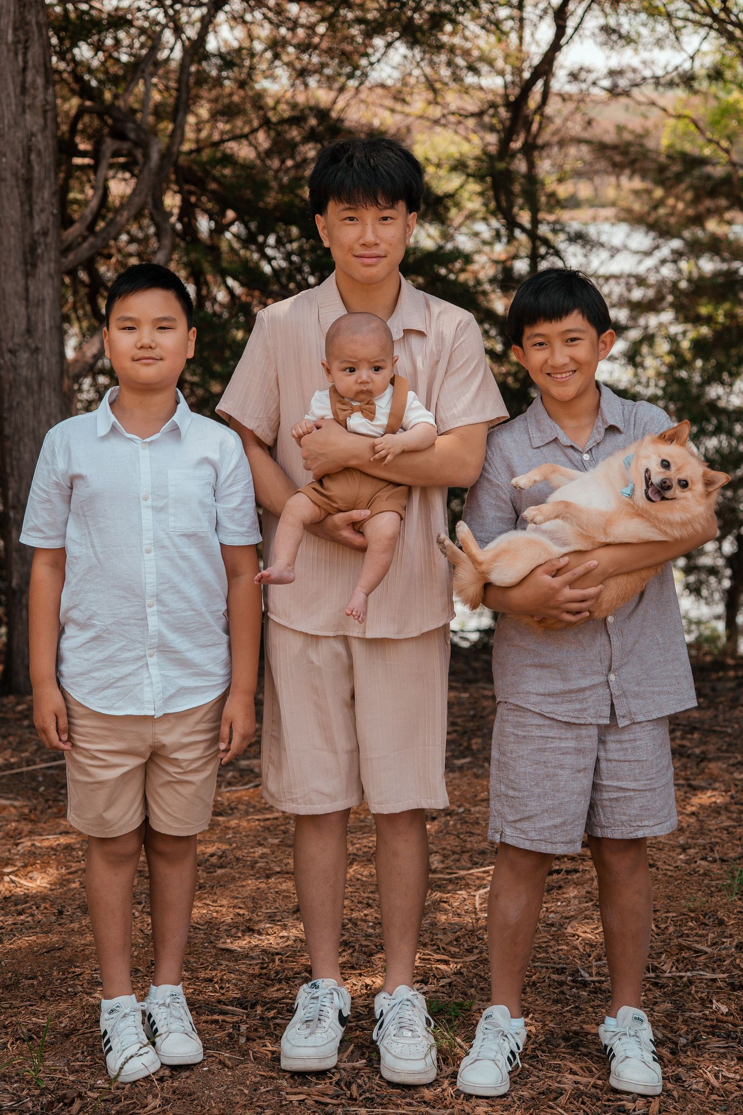 Three people in casual attire stand together outdoors with a baby and a small dog under trees.