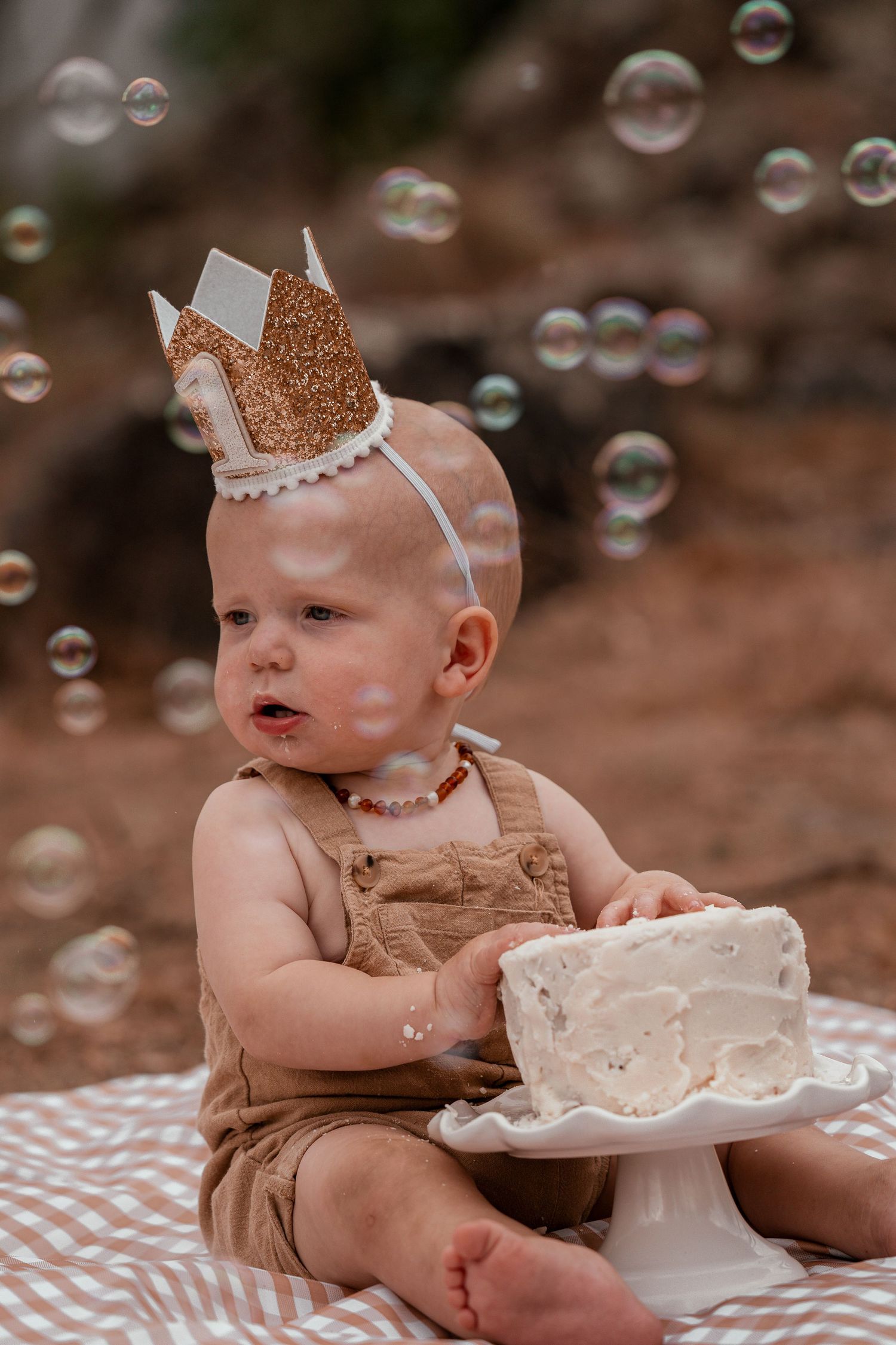 A small child wearing a glittery crown sits on a blanket with a white cake while soap bubbles float in the air.