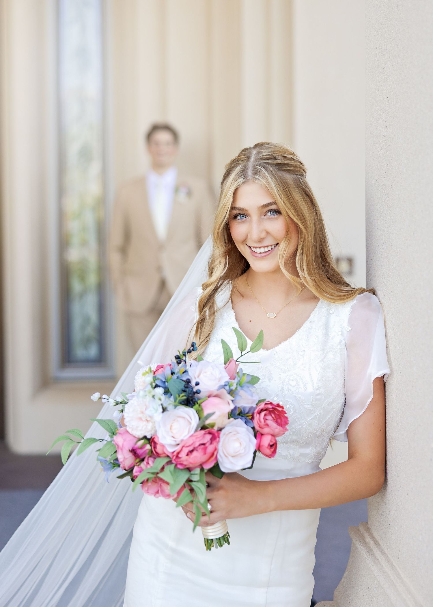 A bride in a white dress smiles while holding a pink and white flower bouquet with a person visible in the background.
