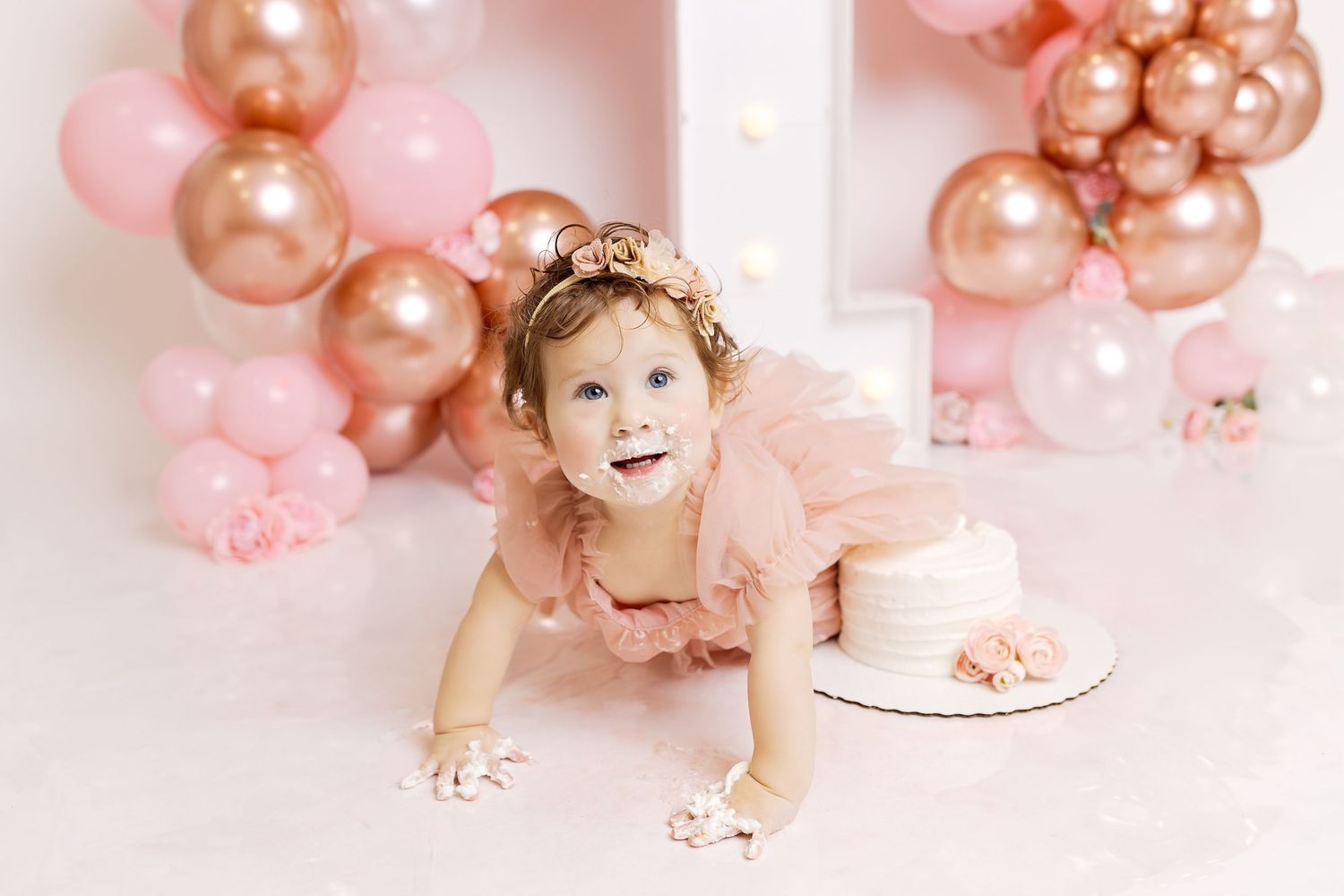 A pink and rose gold balloon arch decorates a birthday cake smash photo session setup with floral accents.