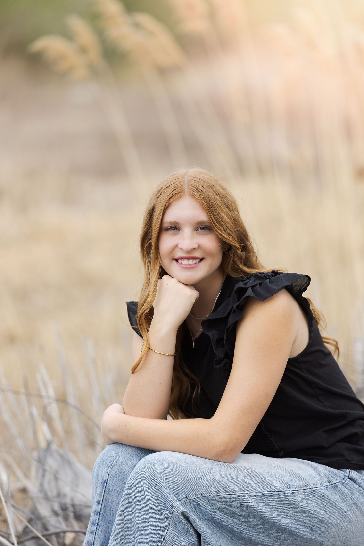 Smiling teenage girl with red hair in black top and jeans sitting outdoors in golden grass field.