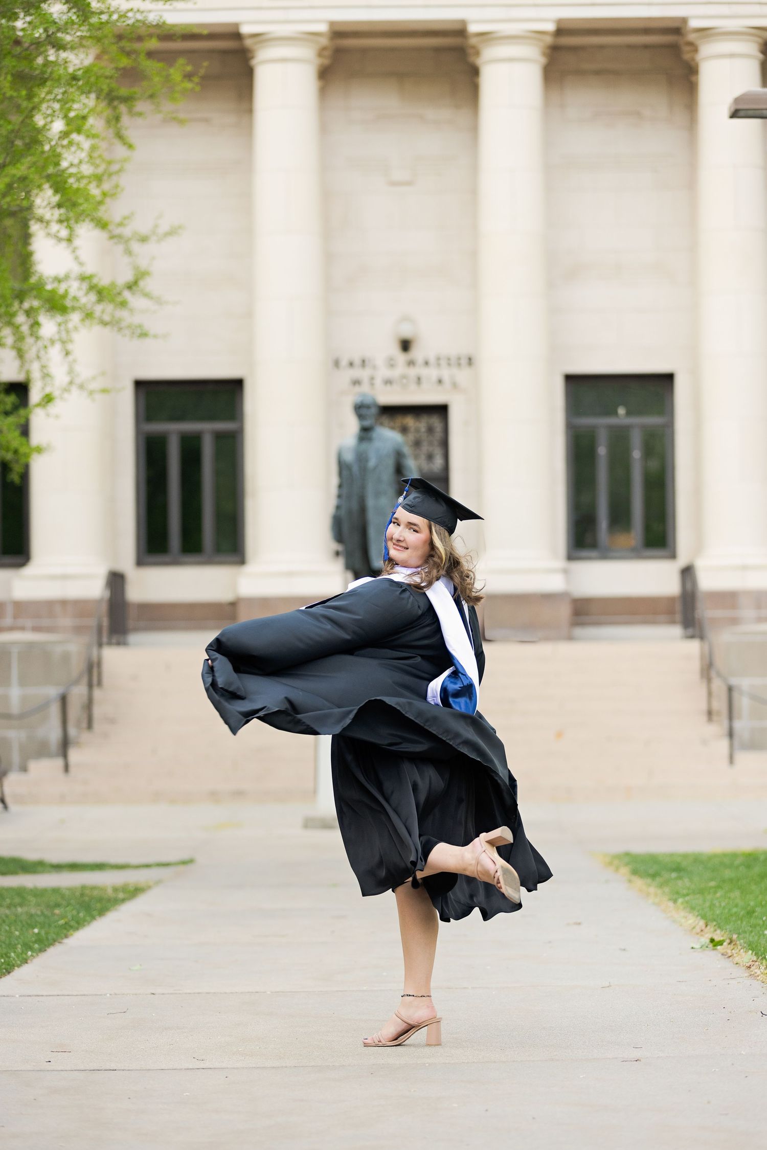 A joyful graduate twirls in black cap and gown on a university campus pathway with a memorial building behind her.