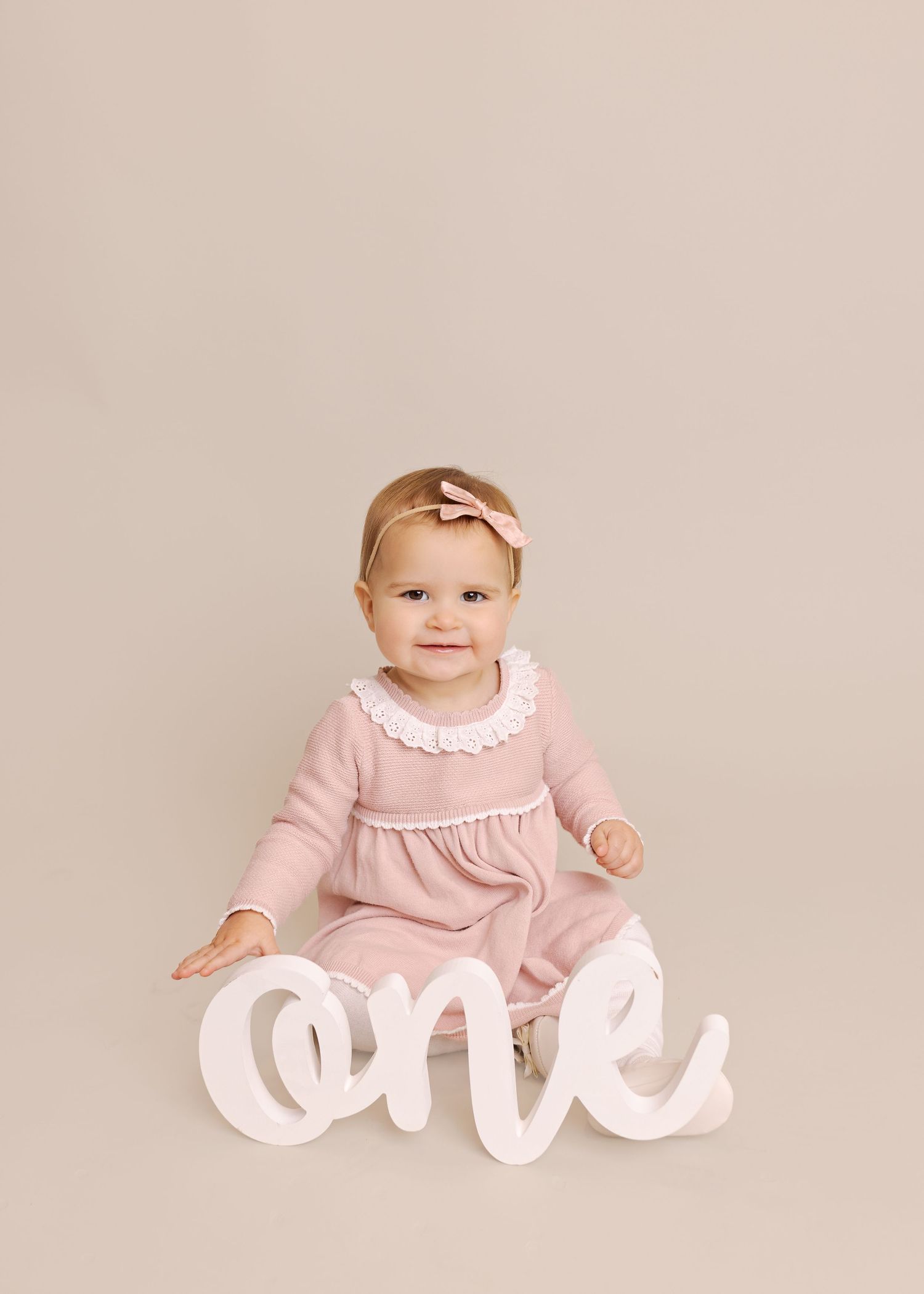 Baby wearing pink dress sits next to white decorative letters spelling 'one' against neutral background.