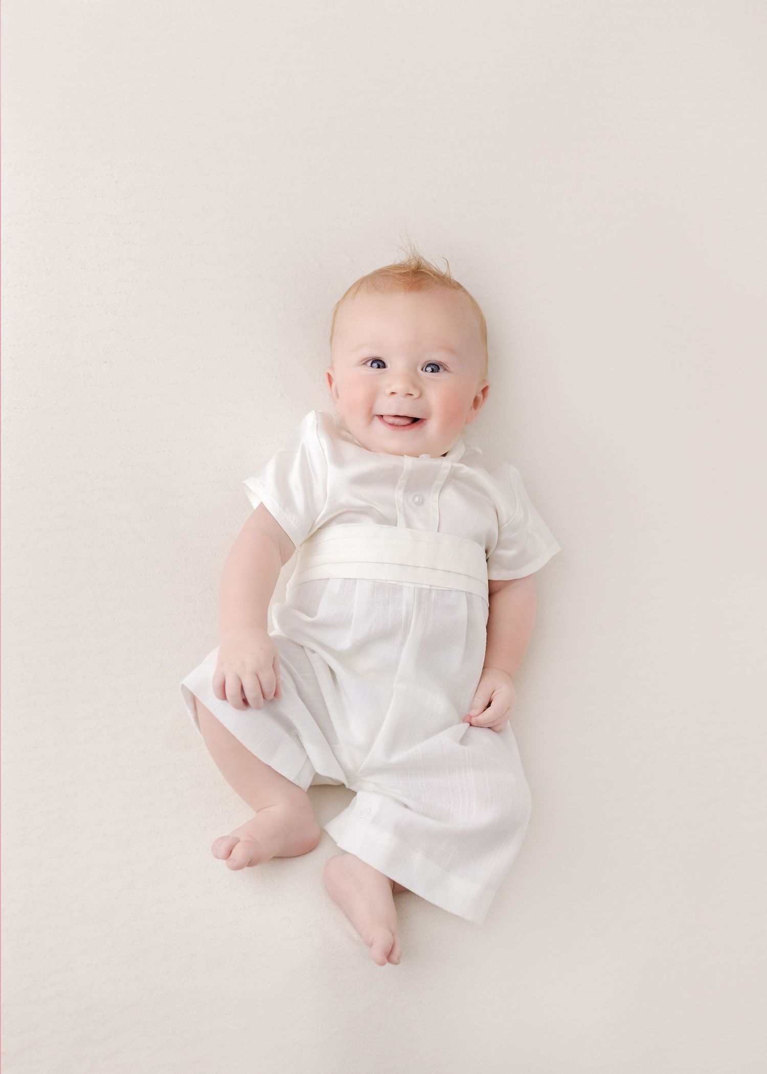 Baby wearing white christening gown lying against neutral background.