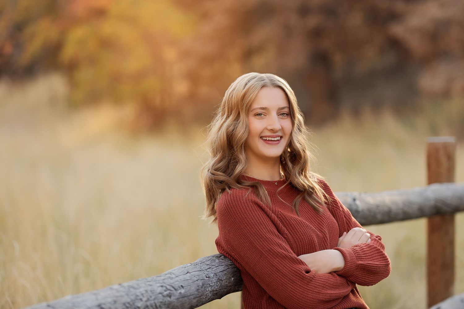 Autumn portrait in burgundy sweater leaning against wooden fence in golden field.
