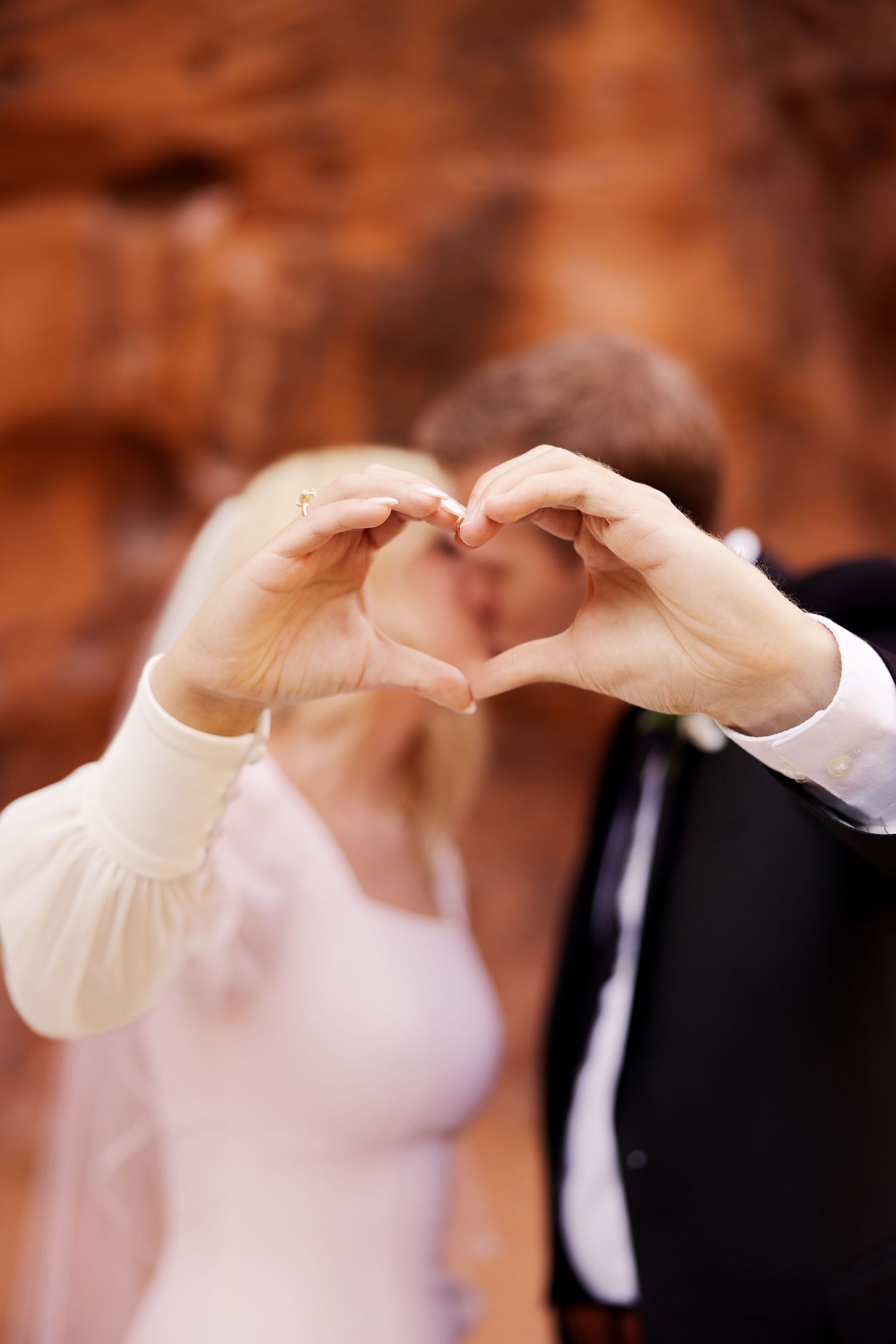 Couple forms heart shape with hands against dramatic red rock backdrop.