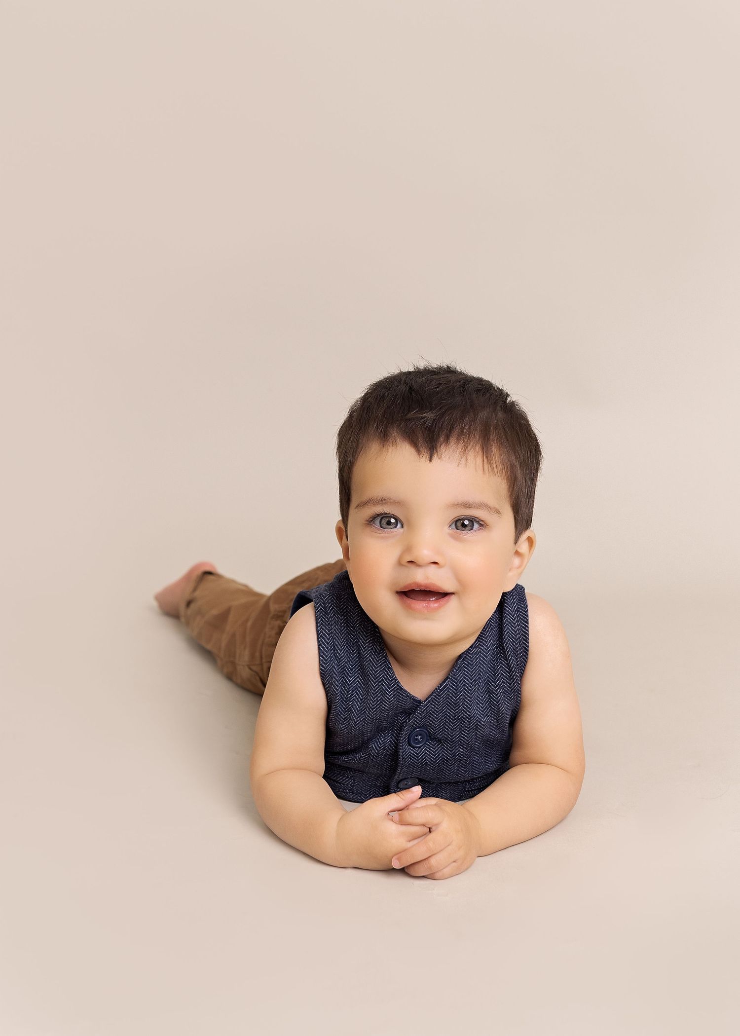 Small child lying on beige backdrop wearing navy blue sleeveless top during professional photo session.