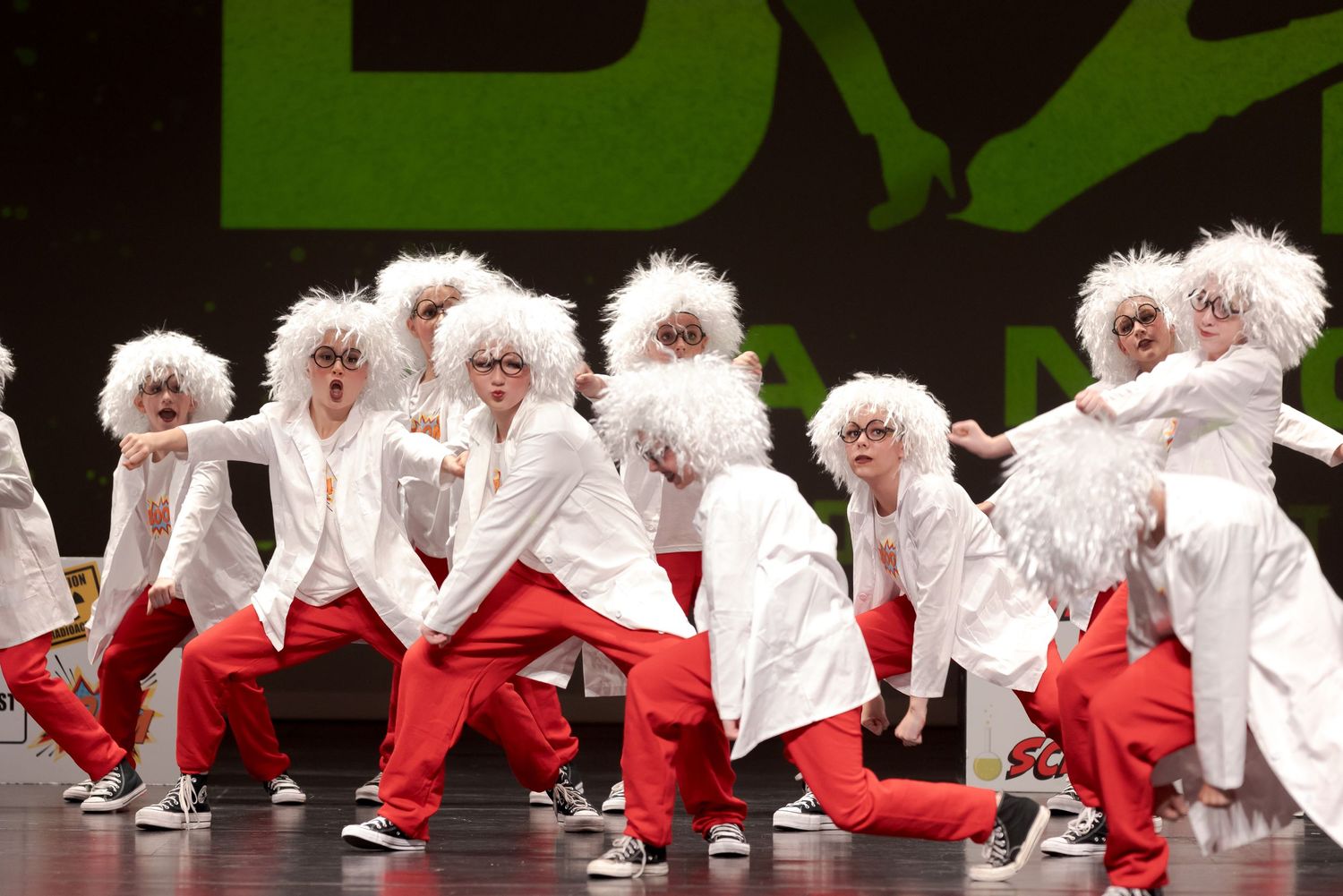 Group of dancers in white furry costumes with red pants perform synchronized dance moves on stage.