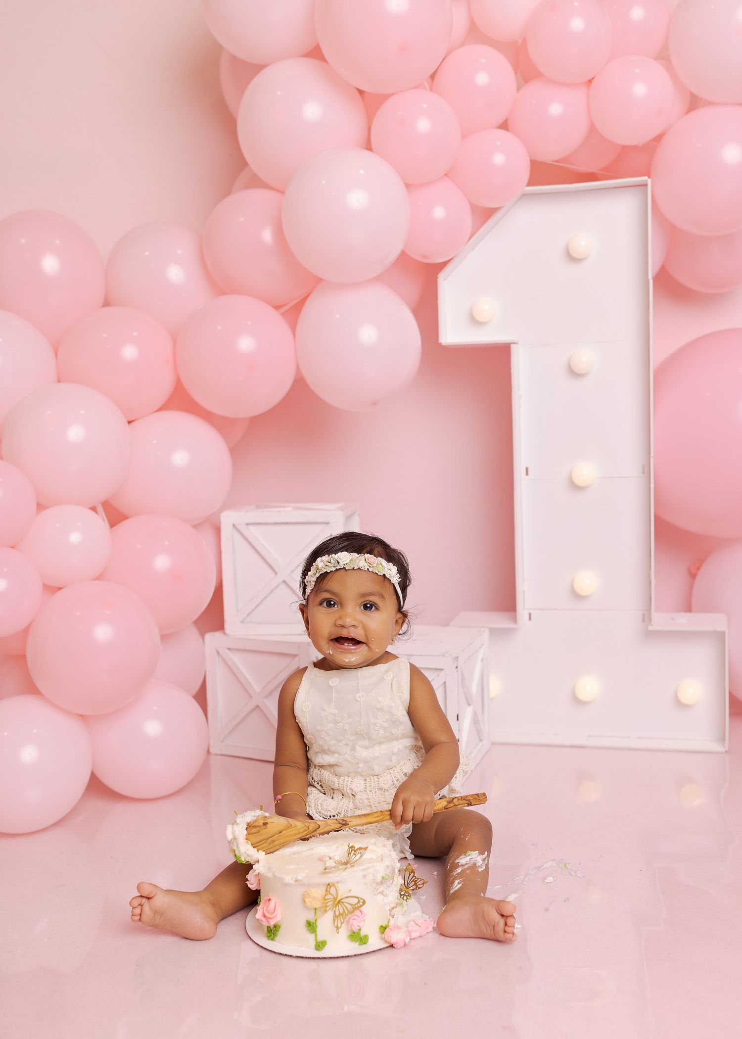 Baby reaches toward birthday cake during cake smash photoshoot with pink balloon background and marquee number one.