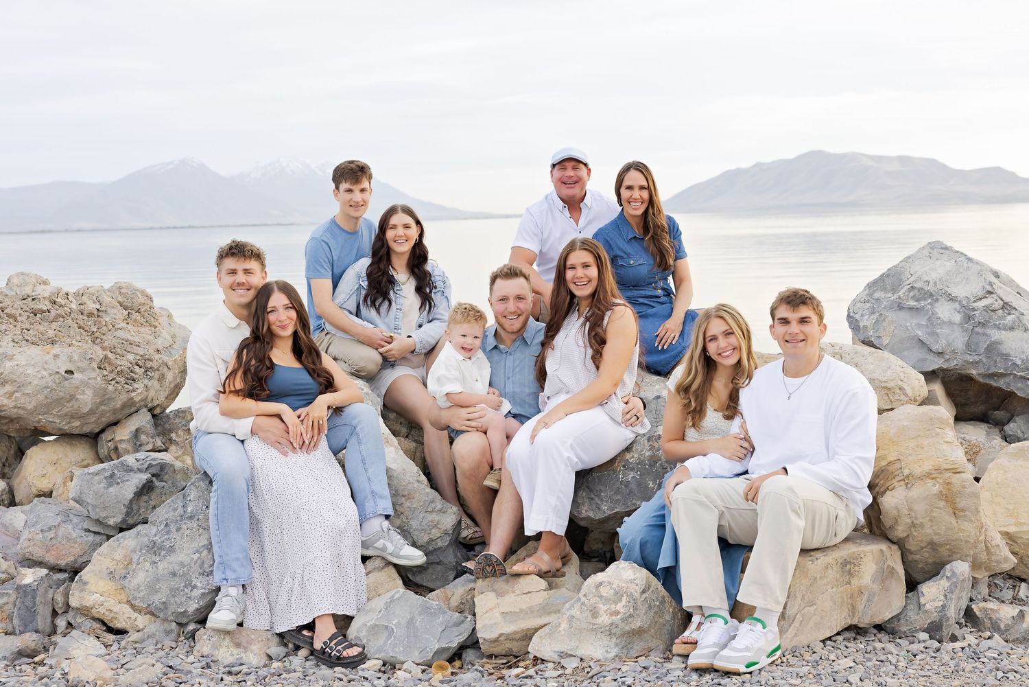 Large family group photo on rocky shoreline with mountain lake backdrop, wearing coordinated white and blue outfits.