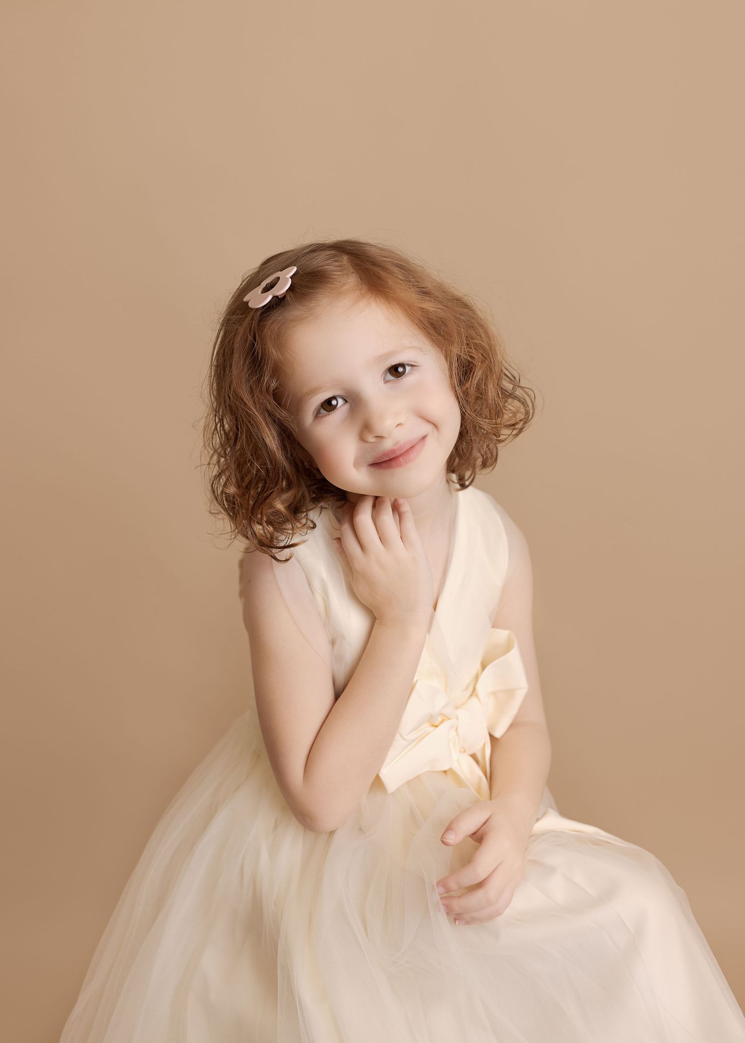 A young girl in a white tulle dress poses sweetly with her hand on her chin.