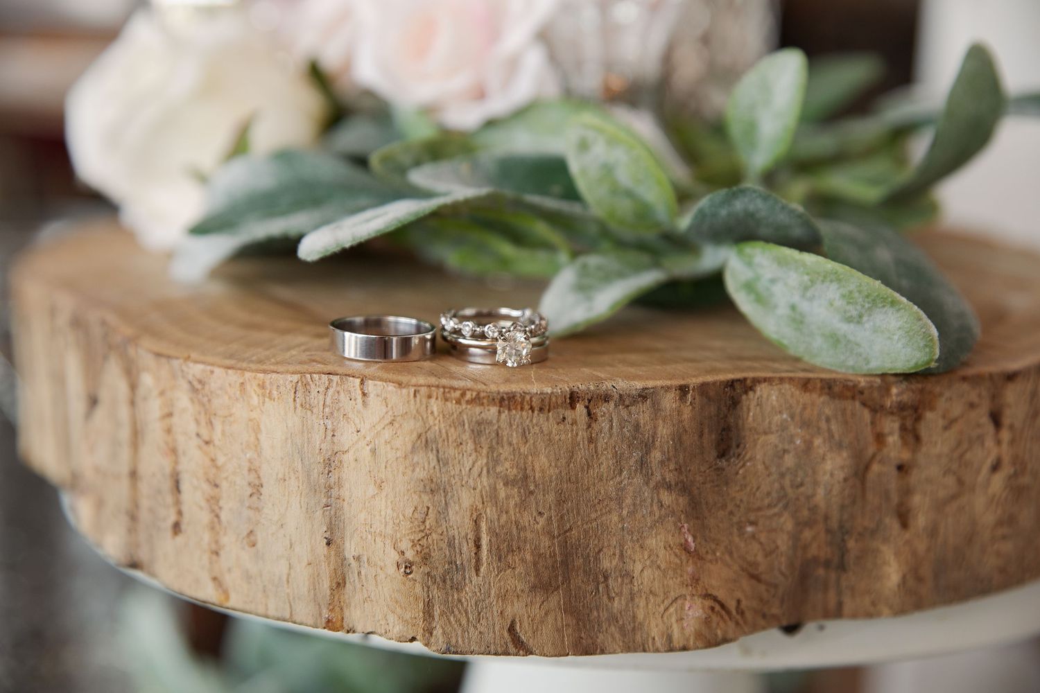 Wedding rings displayed on rustic wood slice with sage leaves and pink flowers in soft focus.