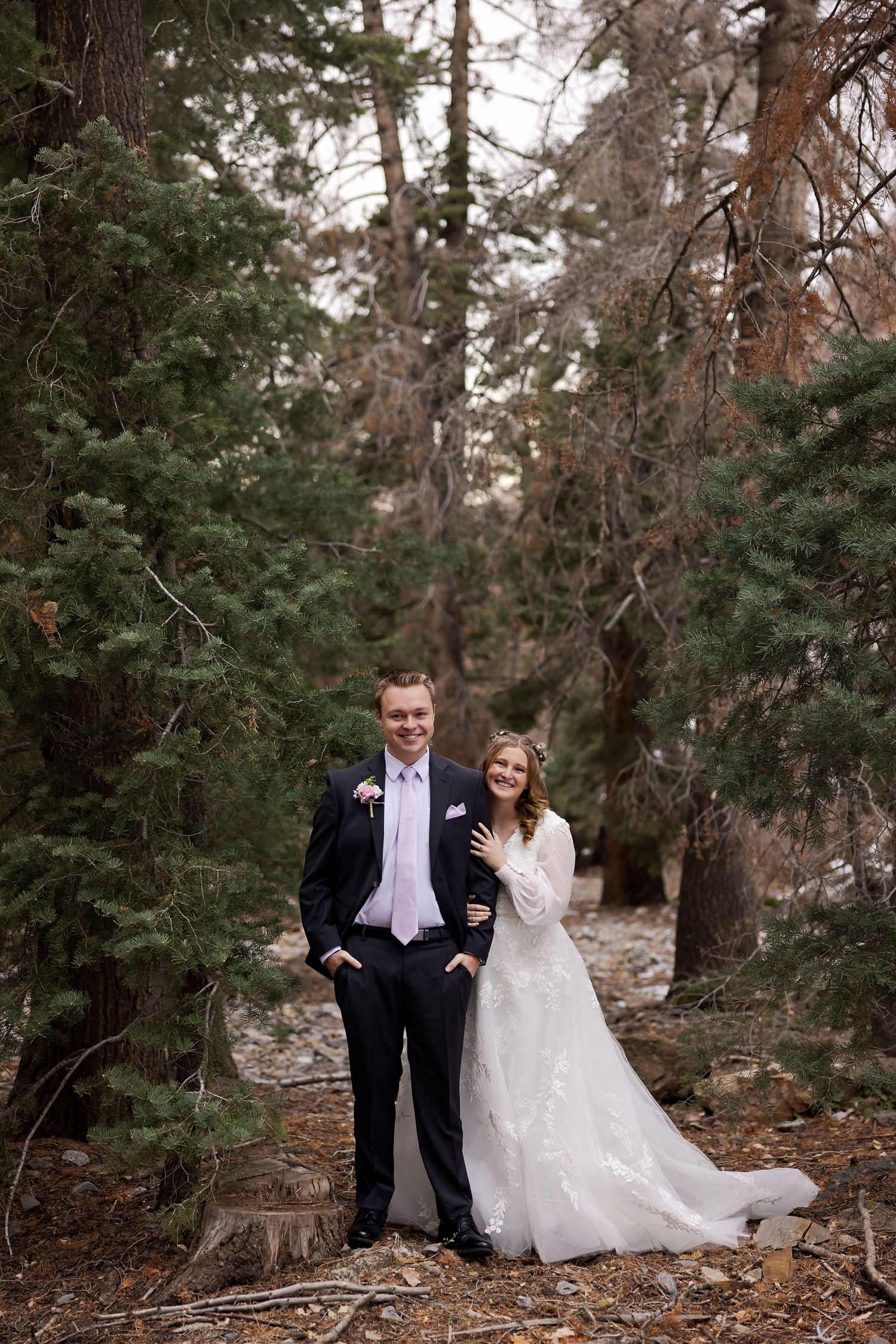 Wedding couple standing among pine trees in rustic forest setting.