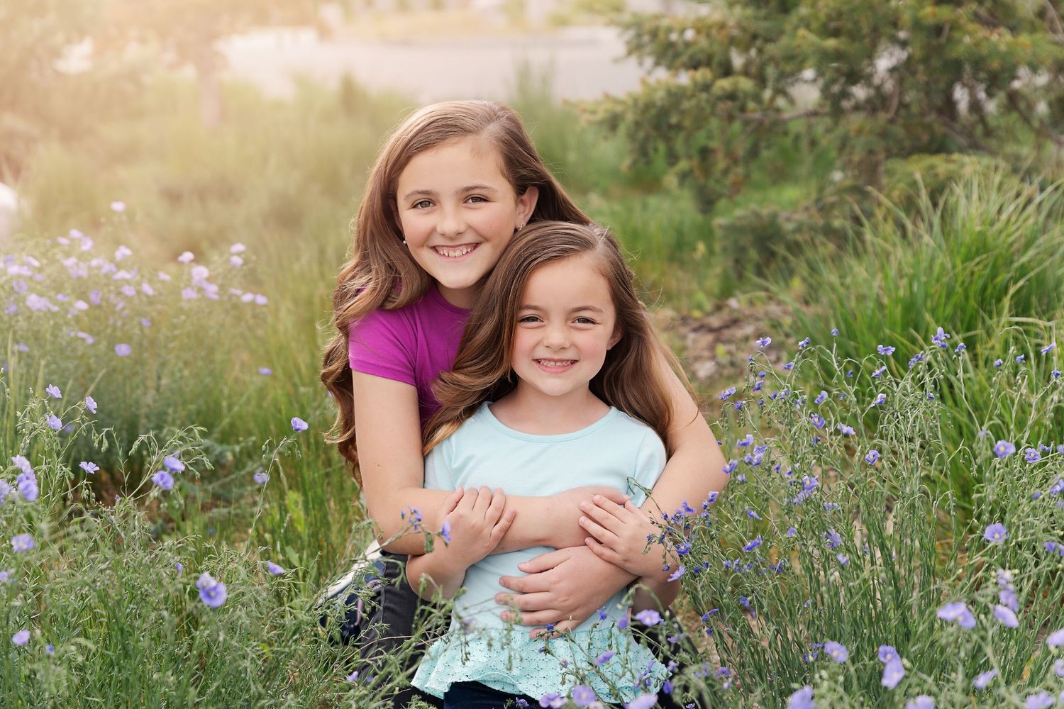 Two young girls embrace and smile while sitting in a field of wildflowers at sunset.