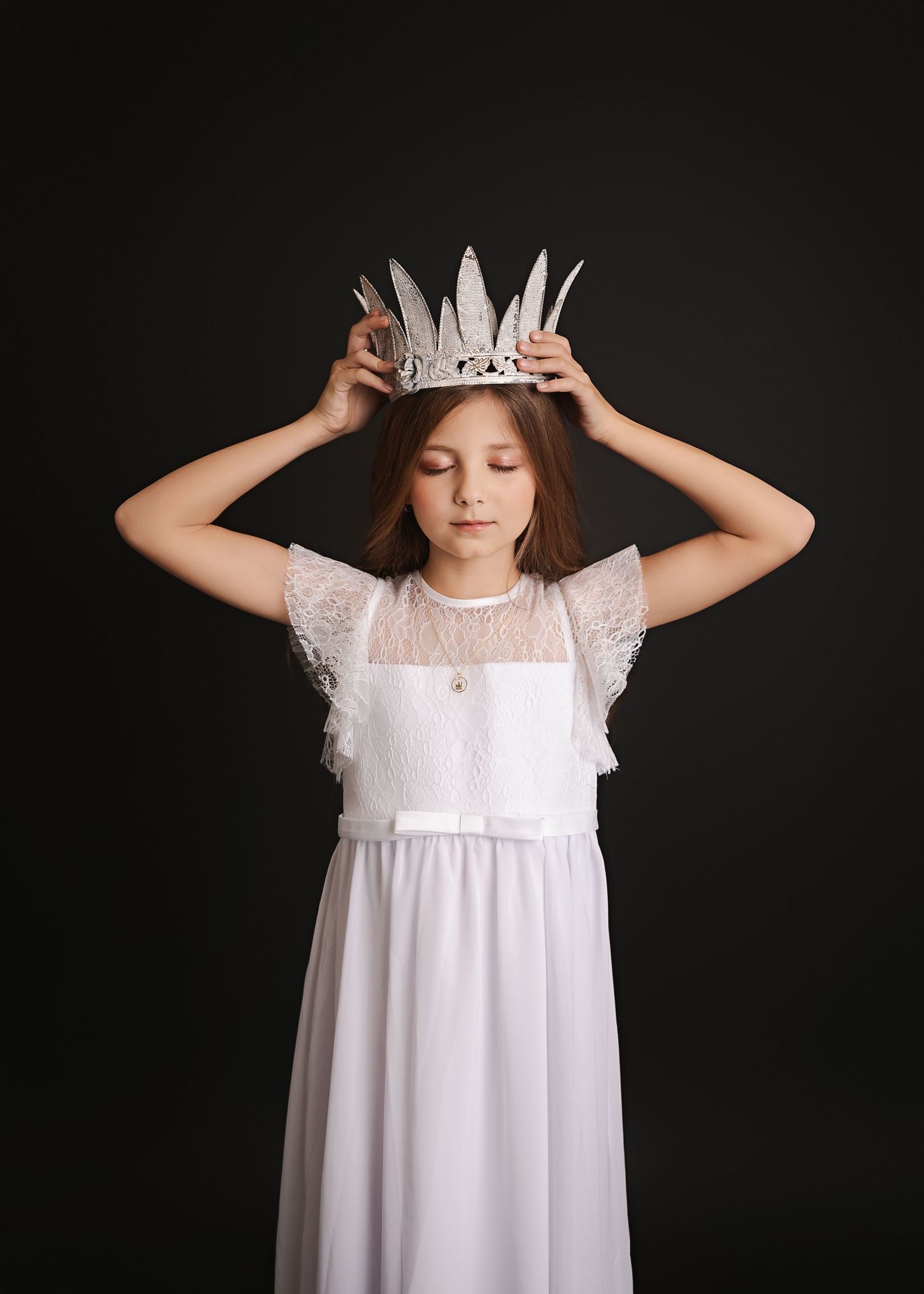 A child in a white lace dress holds a paper crown against a black backdrop.