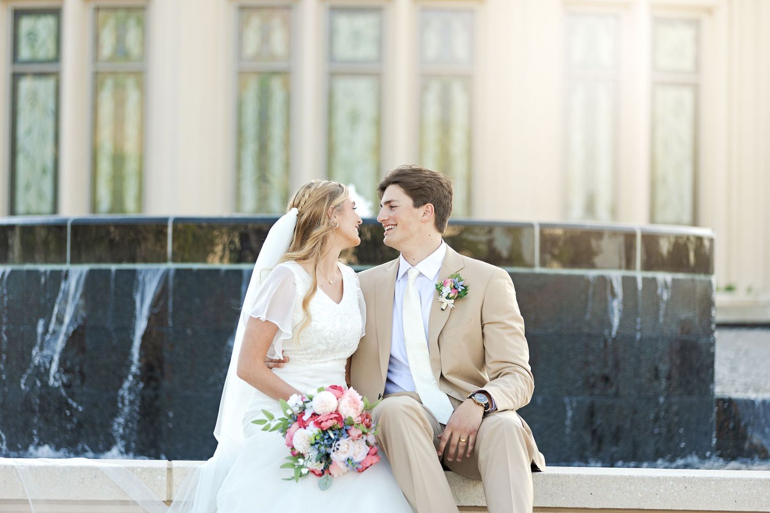 A couple sits together near a fountain sharing a romantic moment in their wedding attire.