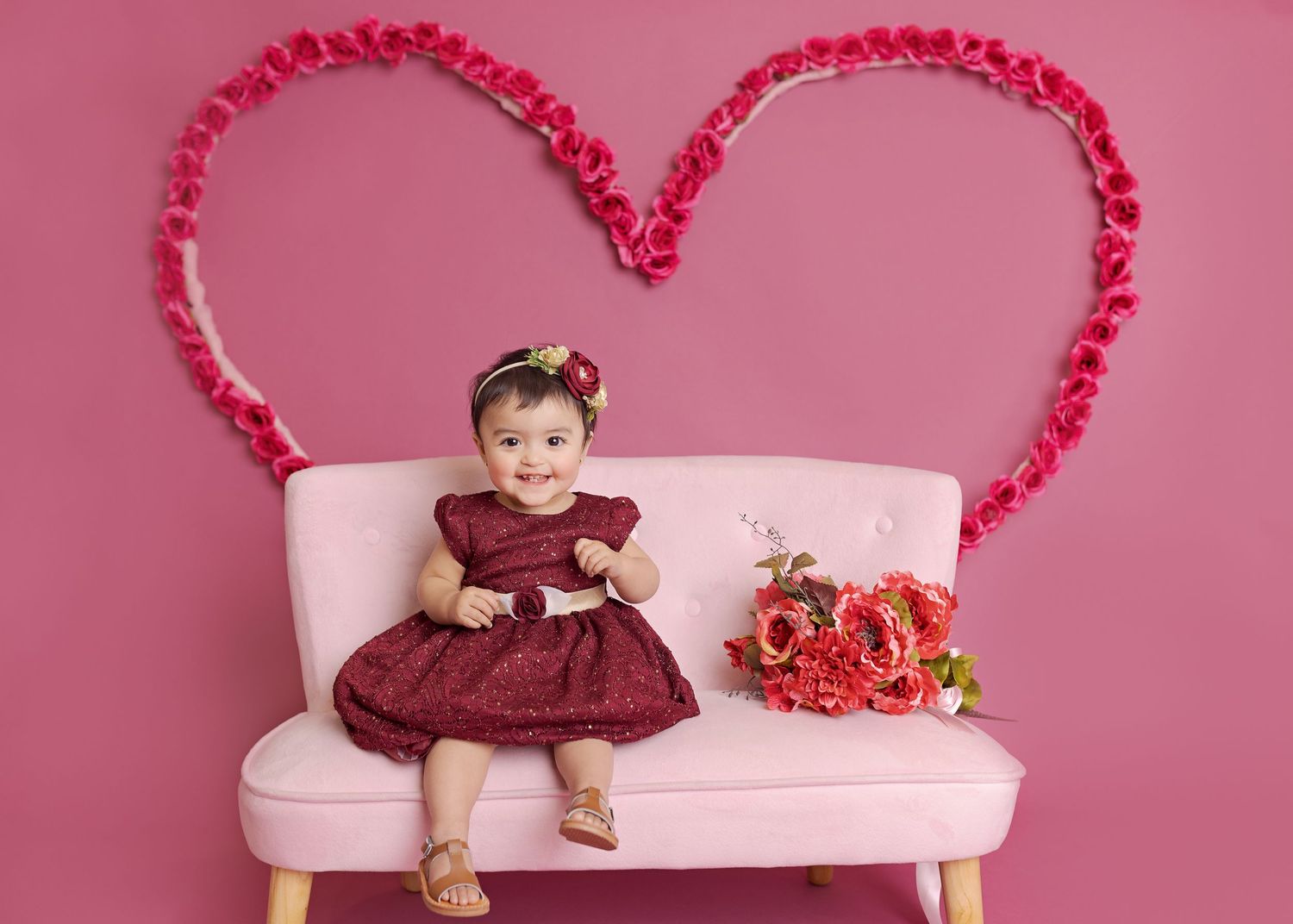 Baby in burgundy dress sits on pink bench with heart decorations and red flowers in Valentine's themed photo shoot.
