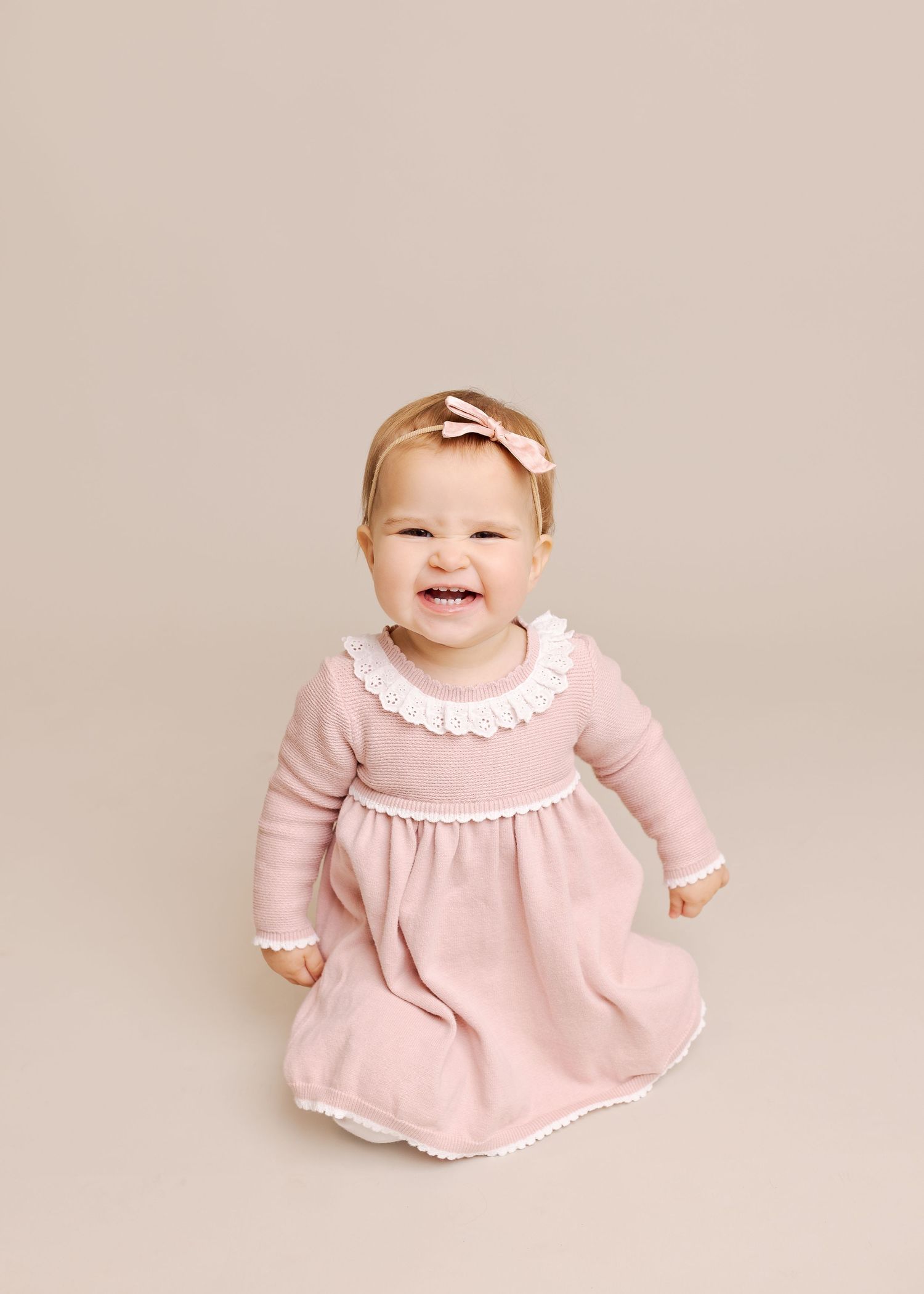 A baby in a pink dress with white trim sits and smiles while crawling across a light beige background.