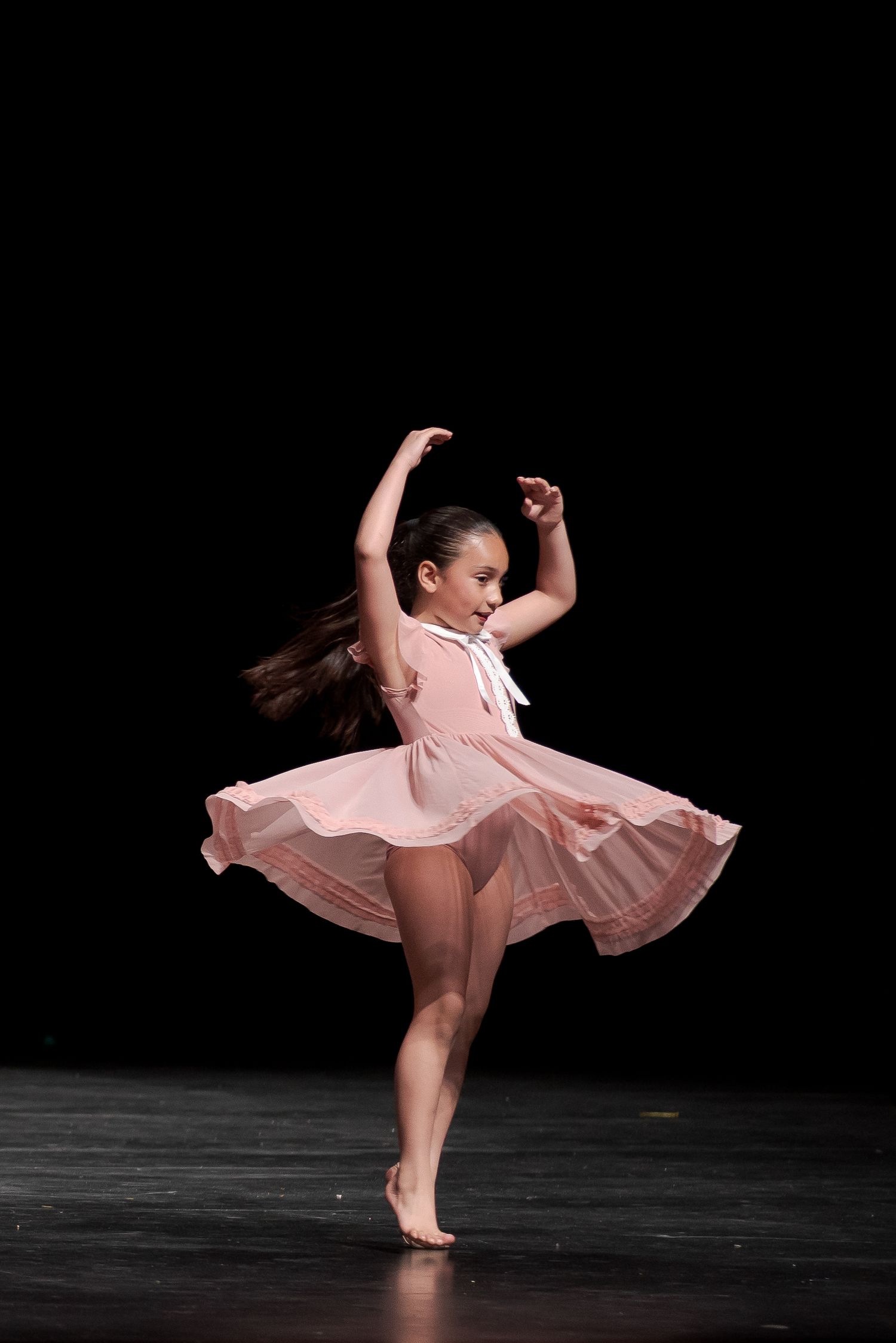 Young dancer in flowing pink dress performs graceful turn on dark stage during dance recital performance.
