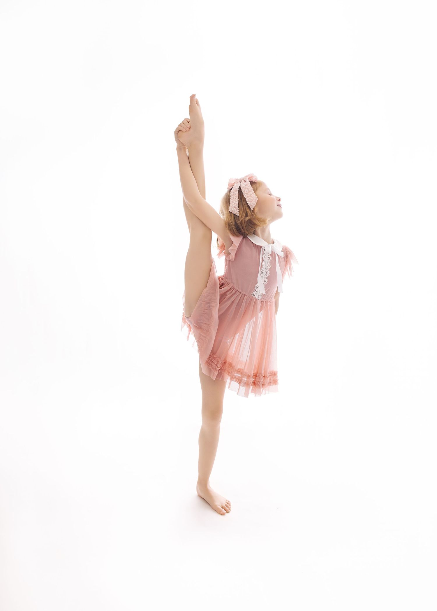 Young dancer in pink dress performs graceful vertical split against white background.